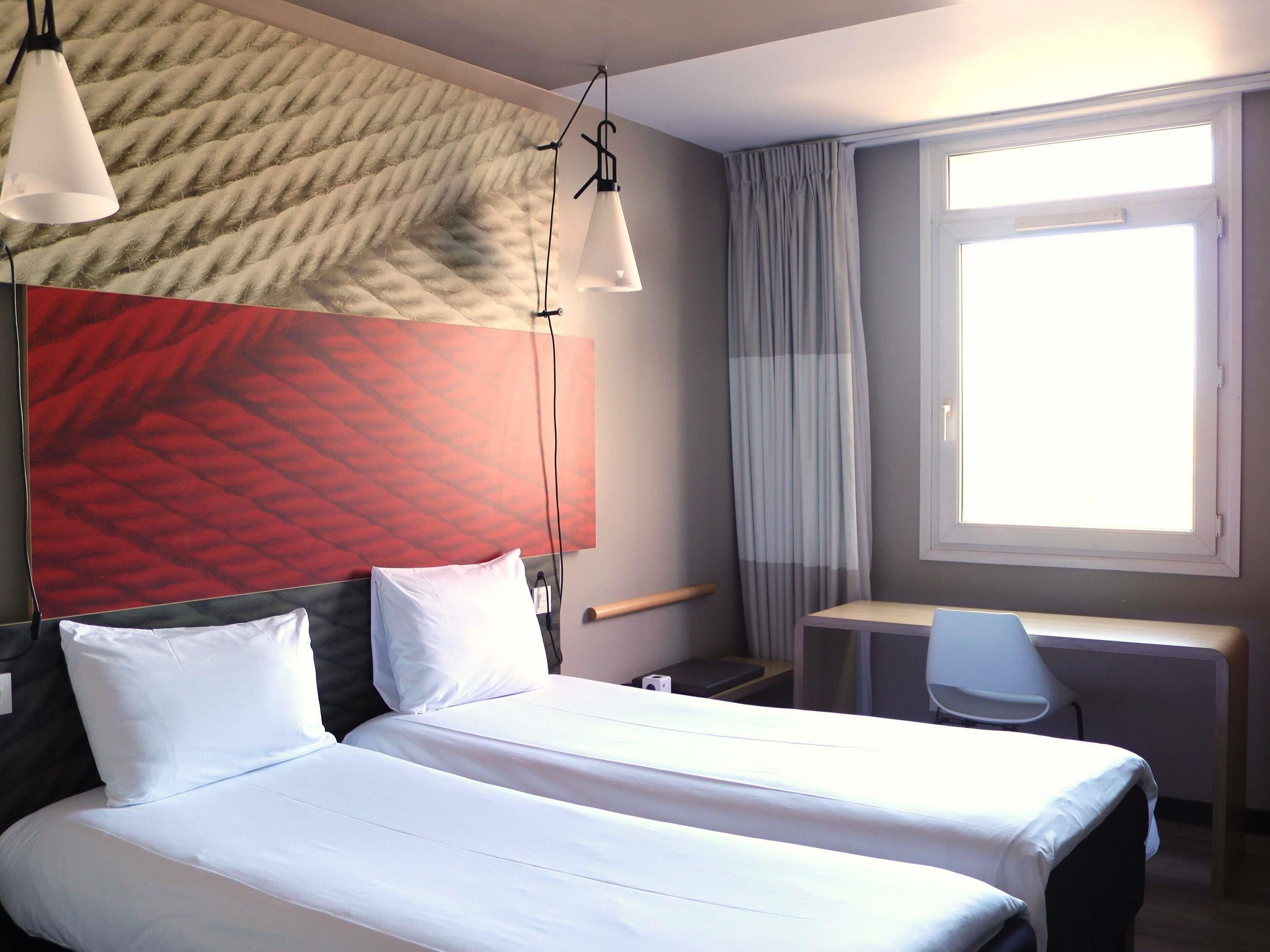 A hotel room with two beds, a desk, a chair, a window with curtains, and a textured wall.