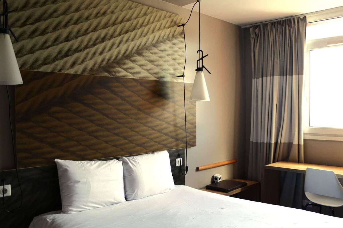 A hotel room with a bed, bedside lamp, and a desk with a chair.