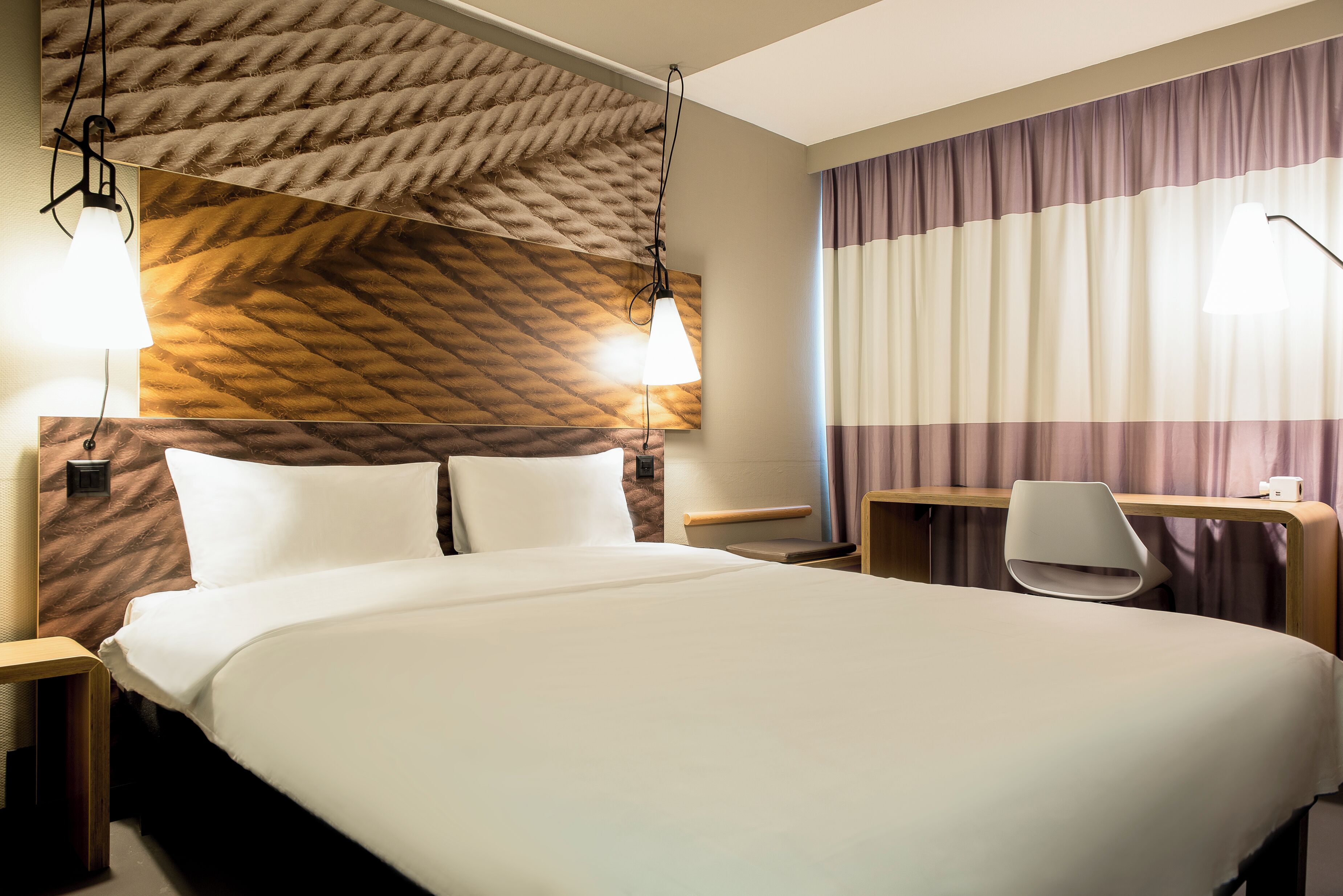 A hotel room with a large bed, two bedside tables, a desk with a chair, and a patterned headboard.