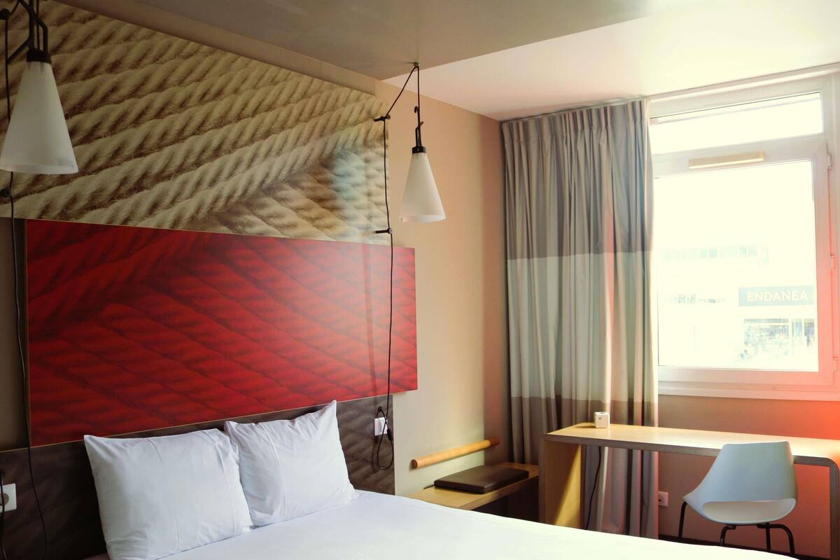 A hotel room with a bed, a desk, a chair, a window with curtains, and a wall with a textured pattern.