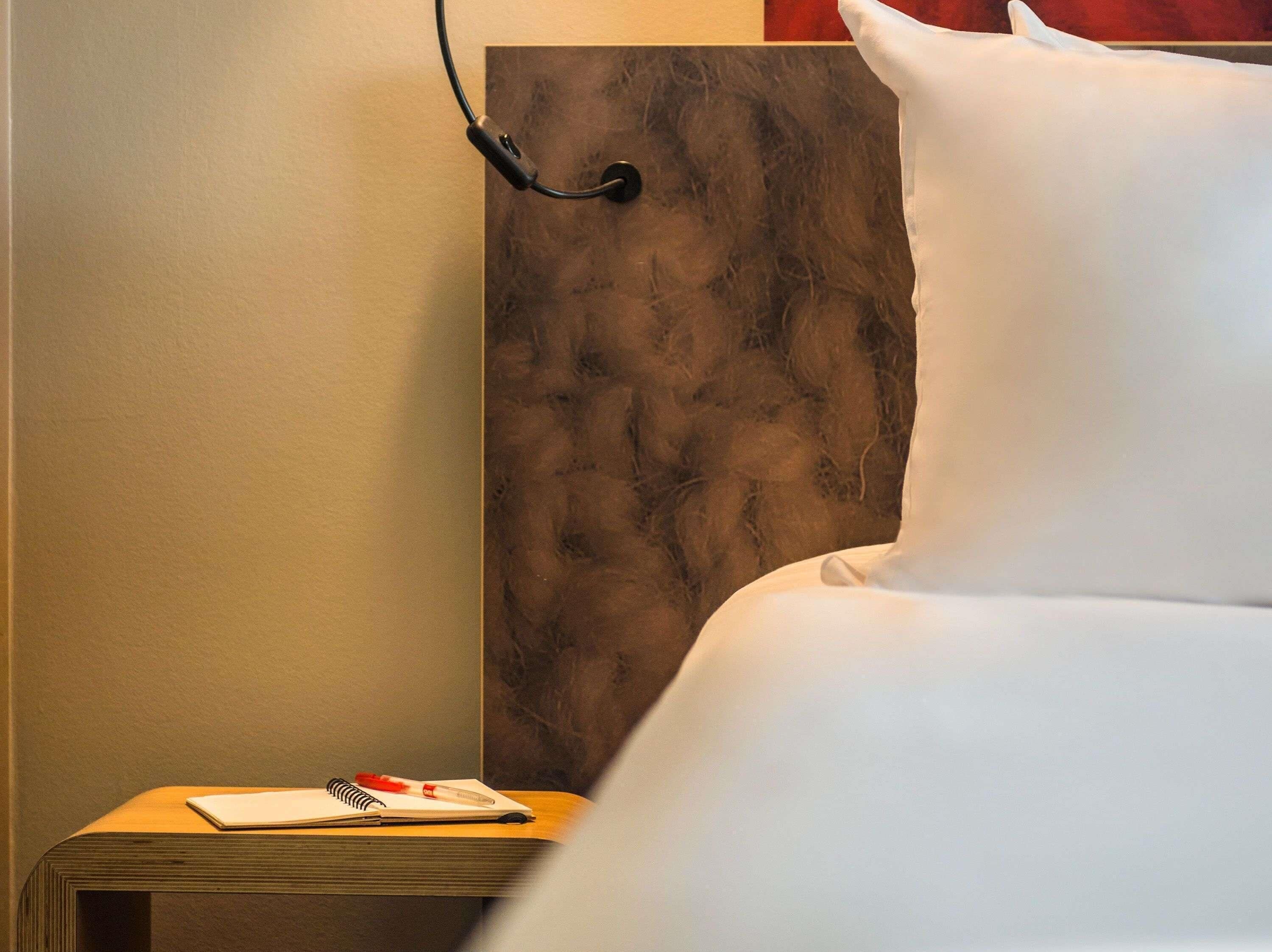 A hotel room with a bed, a nightstand with a notebook and pen, and a wall-mounted lamp.
