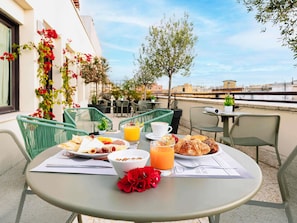 Uma mesa com itens de café da manhã, uma bebida e um cardápio em um terraço com cadeiras e vista para edifícios.