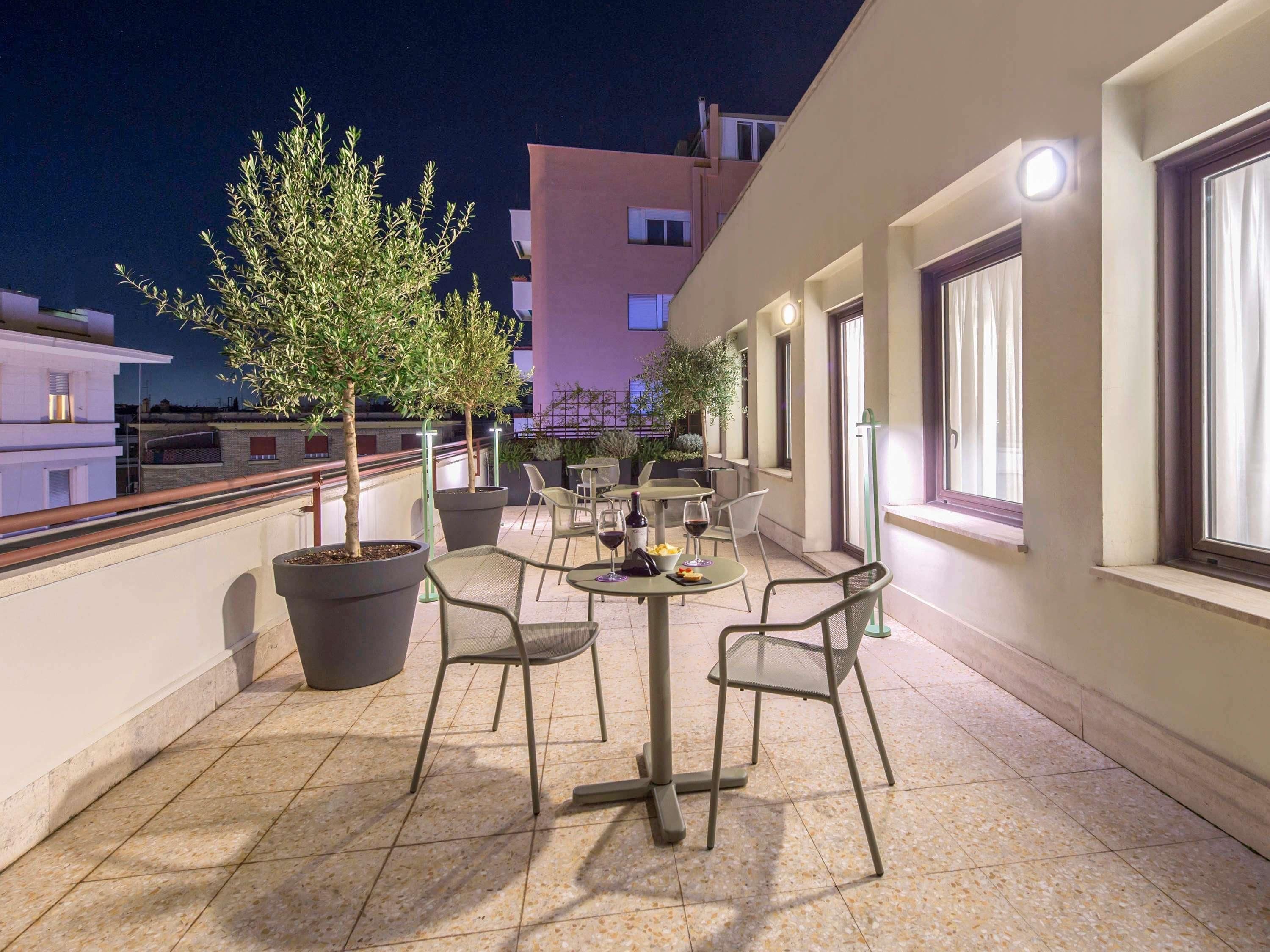 Una terraza al aire libre con árboles en macetas, sillas y mesas, con vistas a un paisaje urbano de noche.
