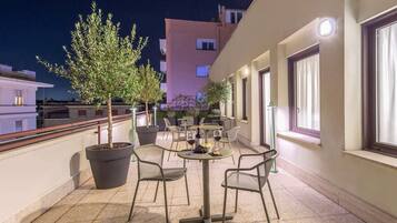 An outdoor terrace with potted trees, chairs, and tables, overlooking a cityscape at night.