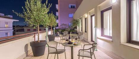 An outdoor terrace with potted trees, chairs, and tables, overlooking a cityscape at night.