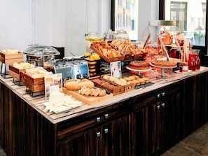 Um buffet com diversos produtos de panificação, incluindo pão, doces e bolos, exposto em um balcão de madeira com vista para uma janela ao fundo.