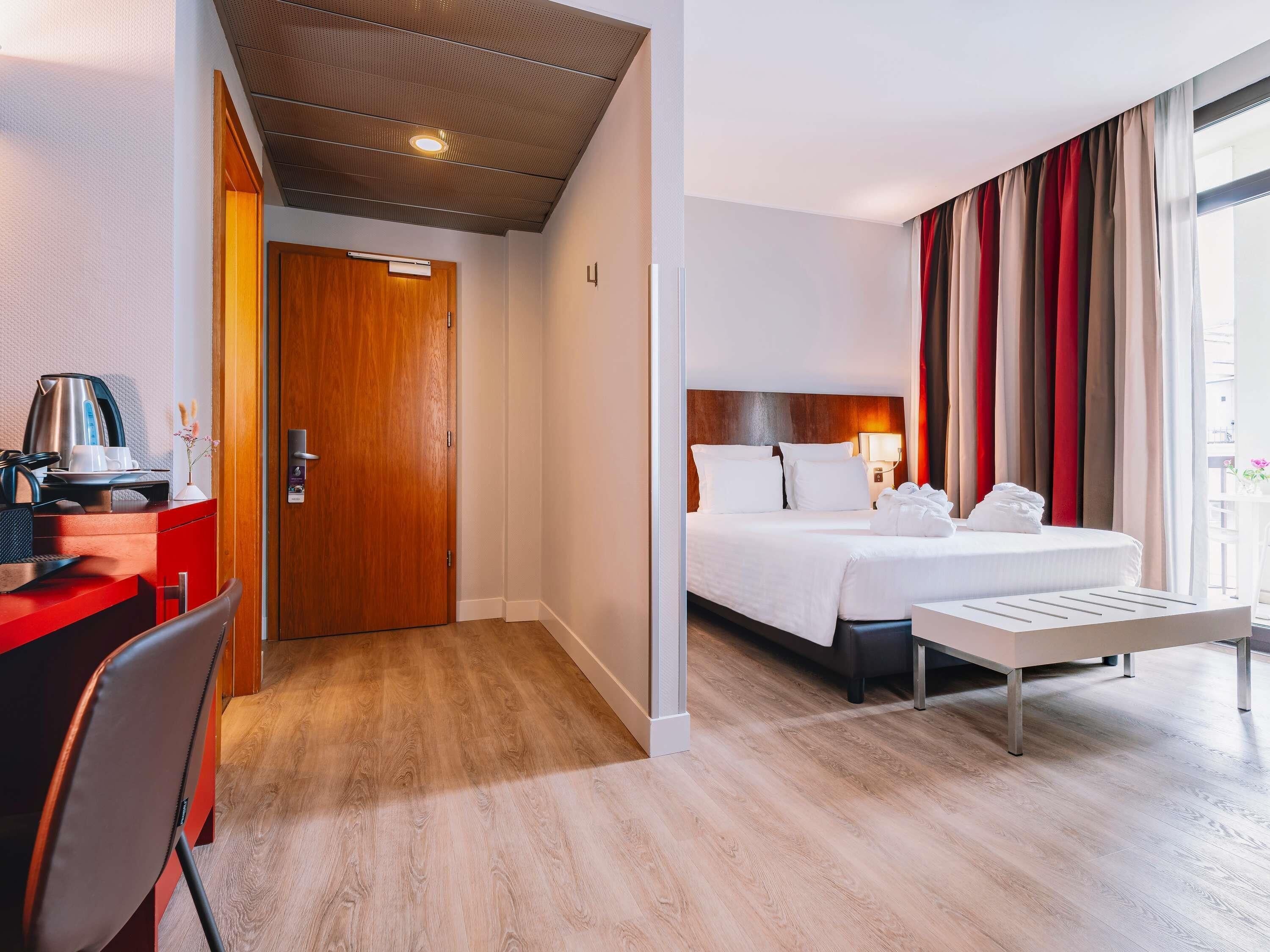 A hotel room with a wooden desk, a bed with white linens, a bedside table, and a window with curtains.