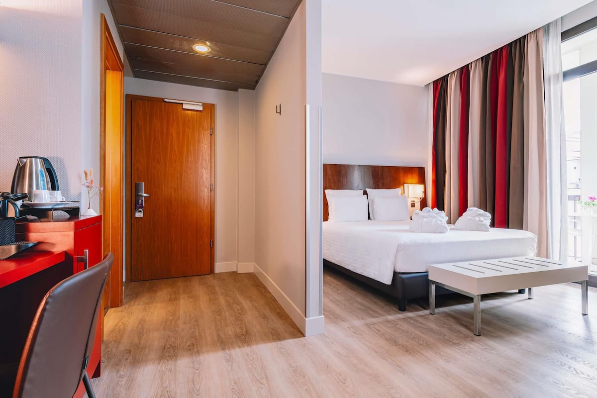 A hotel room with a wooden desk, a bed with white linens, a bedside table, and a window with curtains.