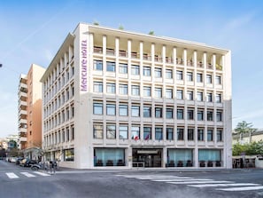 Um hotel de vários andares com um destaque para a placa 'Mercure Hotel', grandes janelas e fachada moderna.