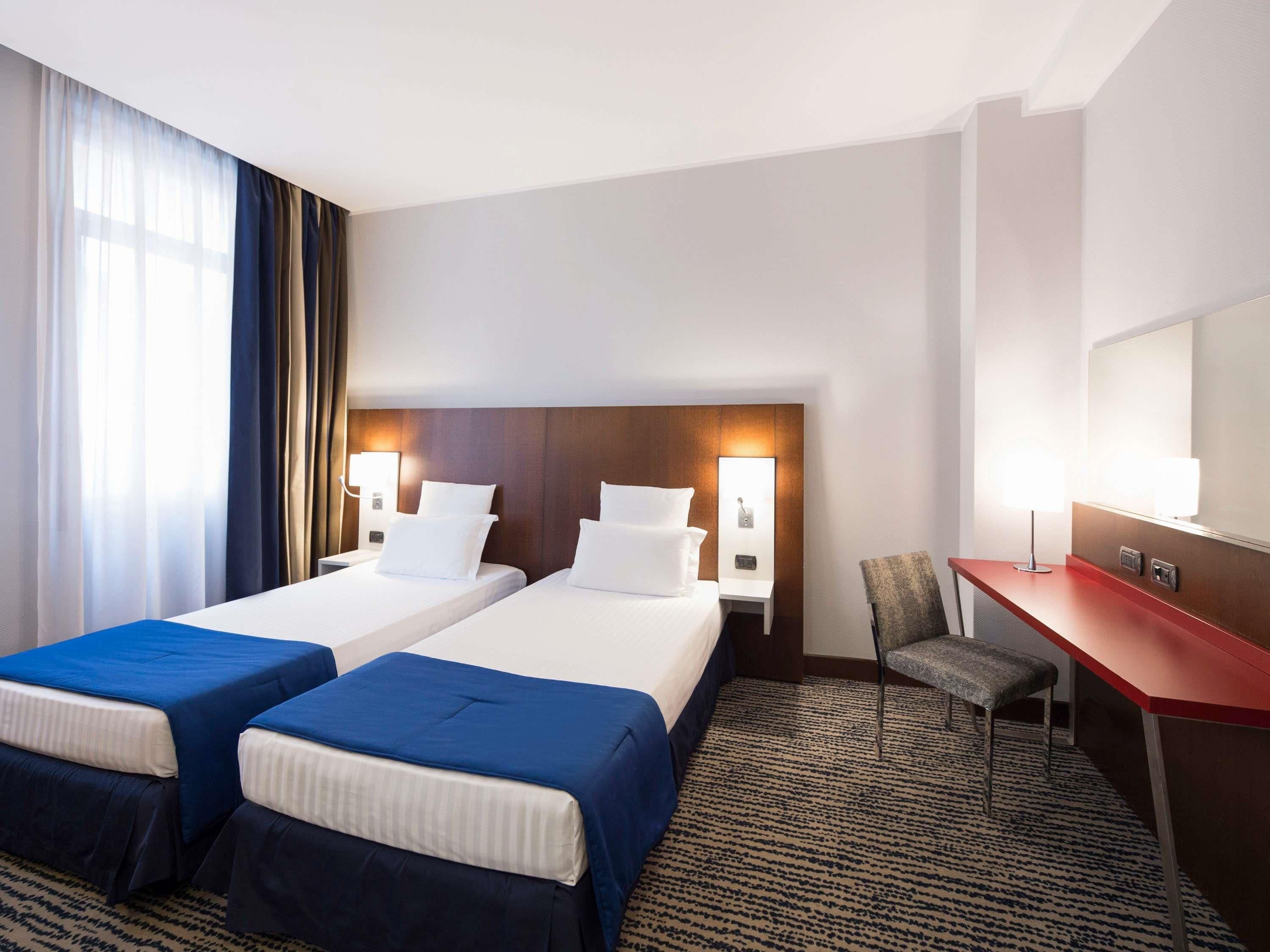 Ein Hotelzimmer mit zwei Betten, einem Schreibtisch, einem Sessel und einem großen Fenster mit Vorhängen.
