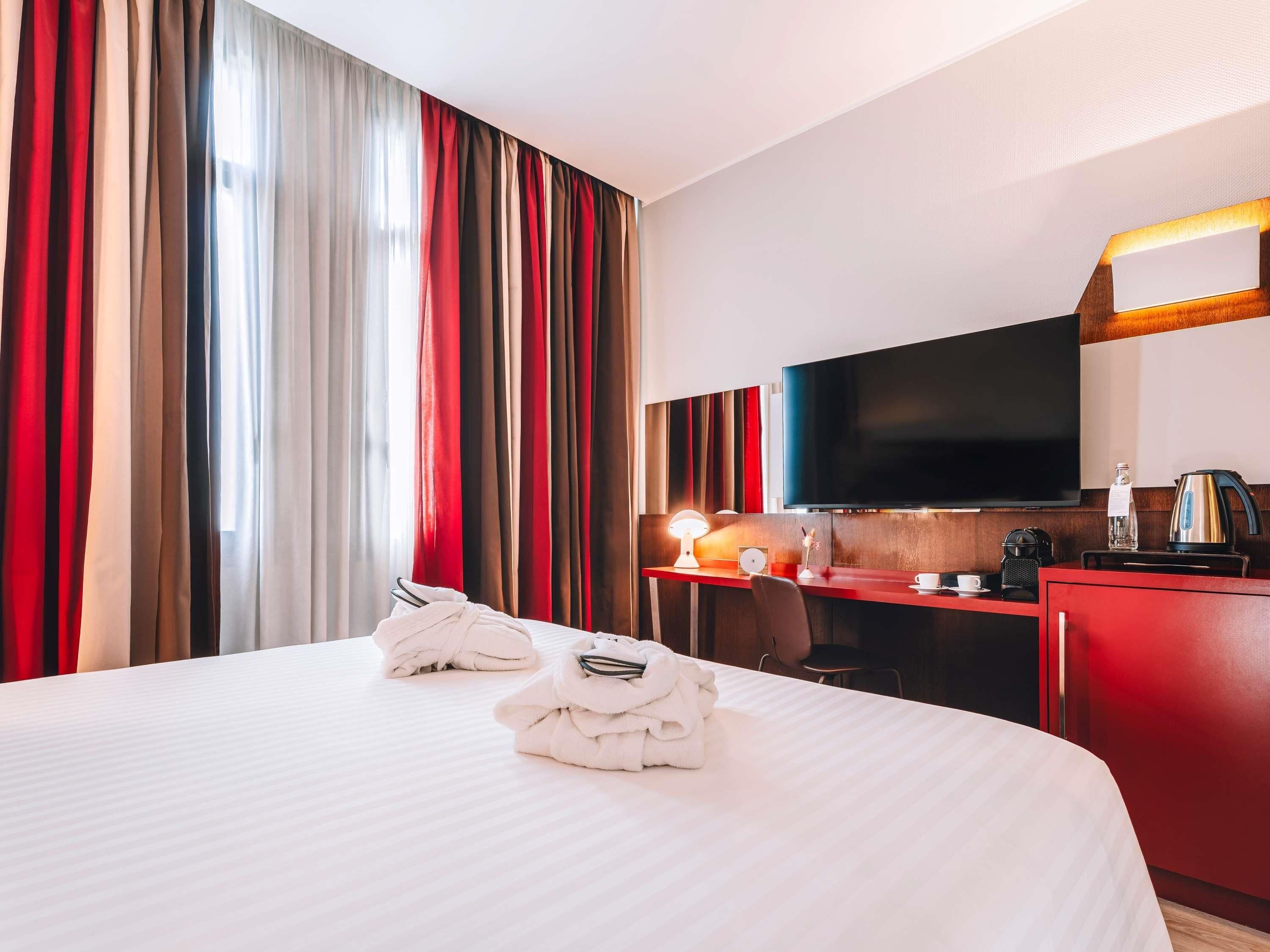 A hotel room with a bed, a desk with a TV, a coffee maker, and a red cabinet.