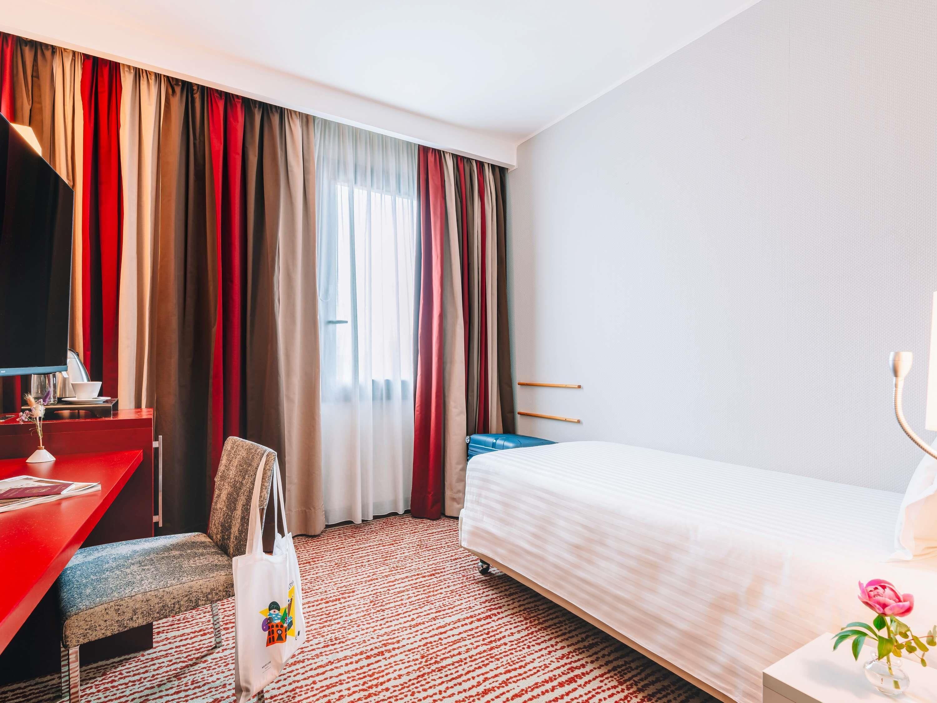 A hotel room with a bed, a desk with a computer, a chair, and a red wall.
