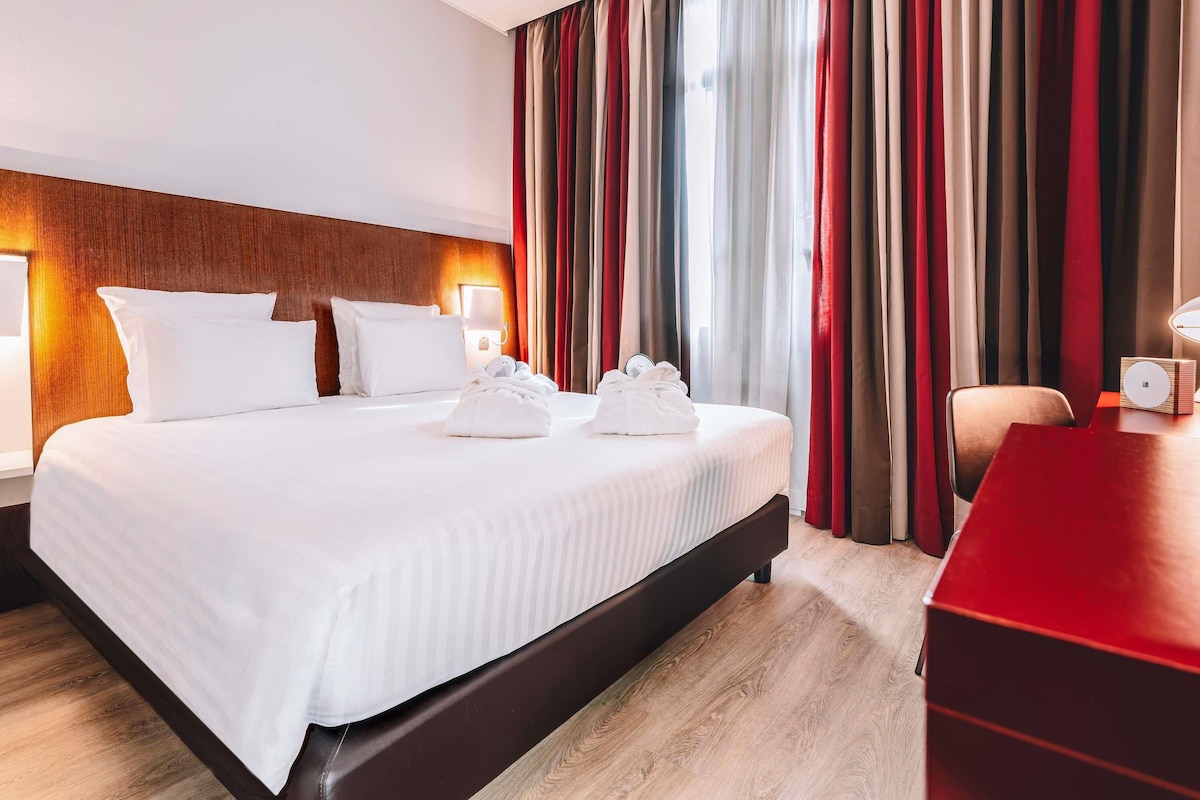 A hotel room with a large bed, a wooden headboard, a desk with a lamp, and a red chair.