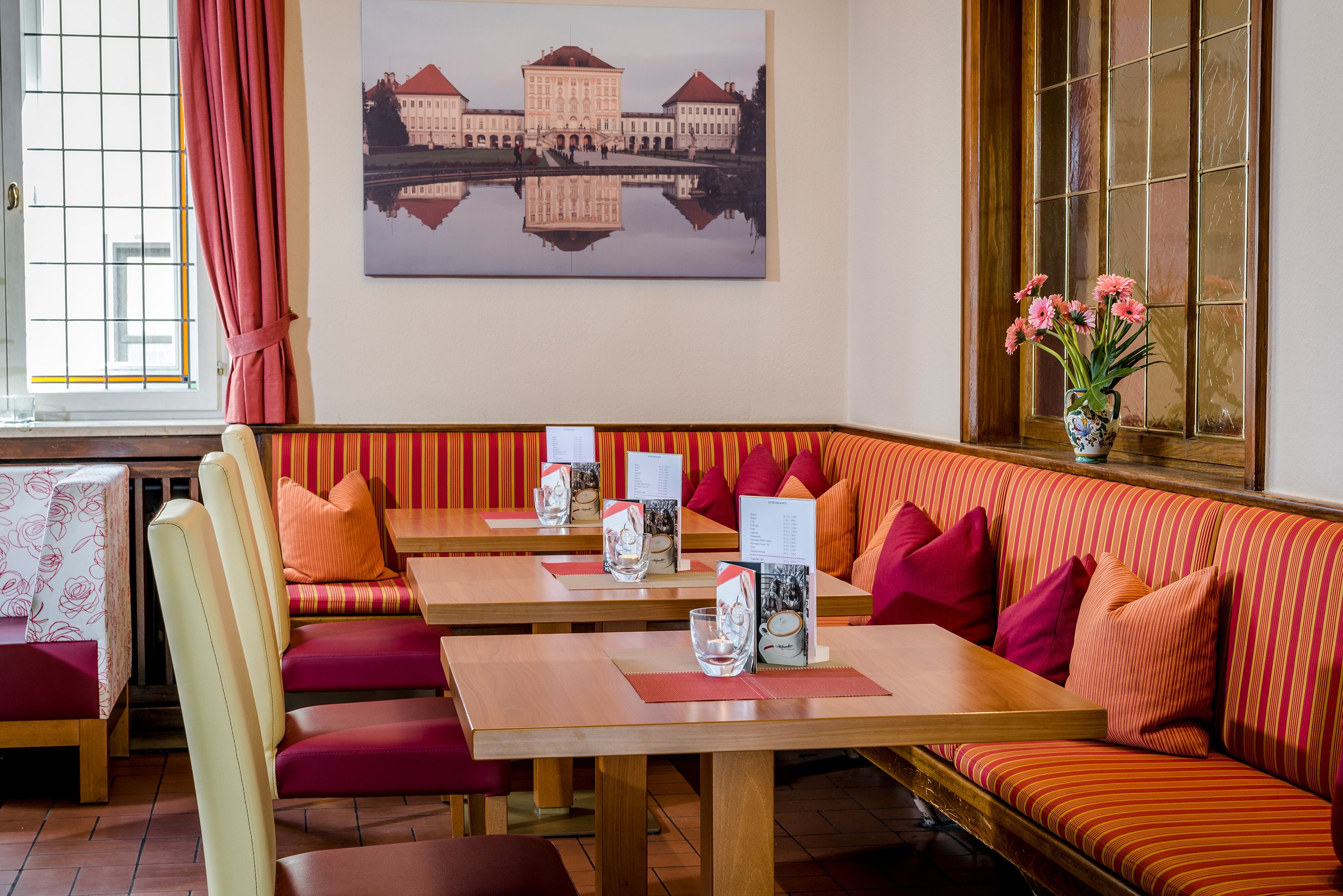 Un restaurante con asientos a rayas rojas y naranjas, mesas de madera y un florero con flores rosas en el alféizar de una ventana.