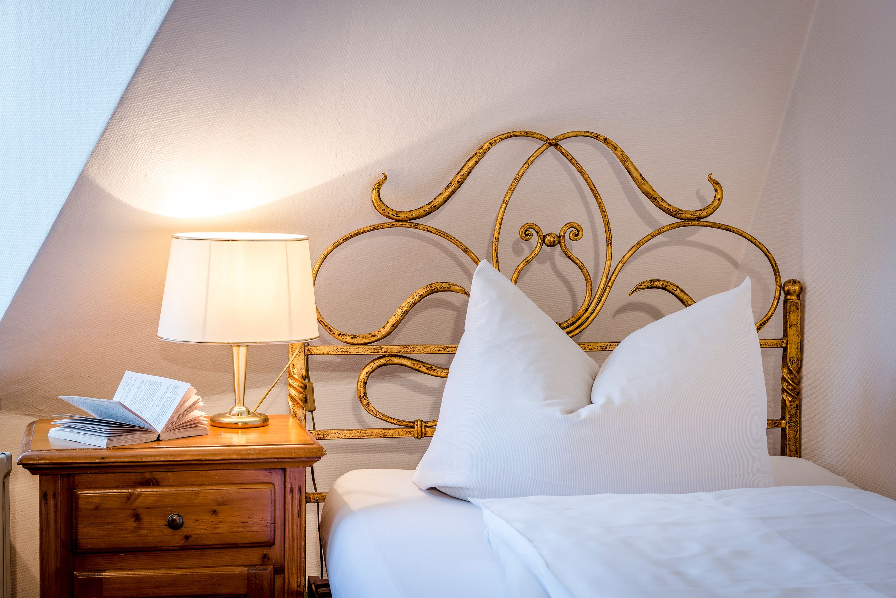 A neatly made bed with white bedding, a gold-framed headboard, and a bedside table with a lamp and an open book.