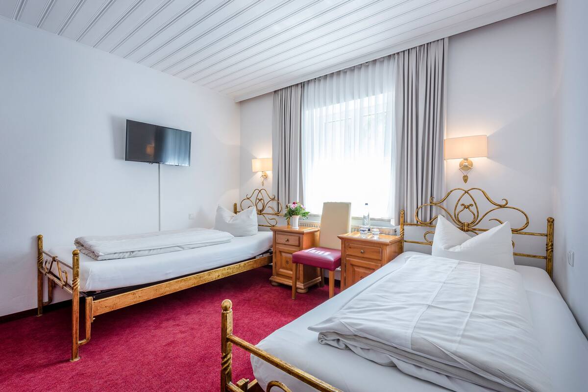 A hotel room with two single beds, a desk with a chair, a TV mounted on the wall, and a large window with curtains.