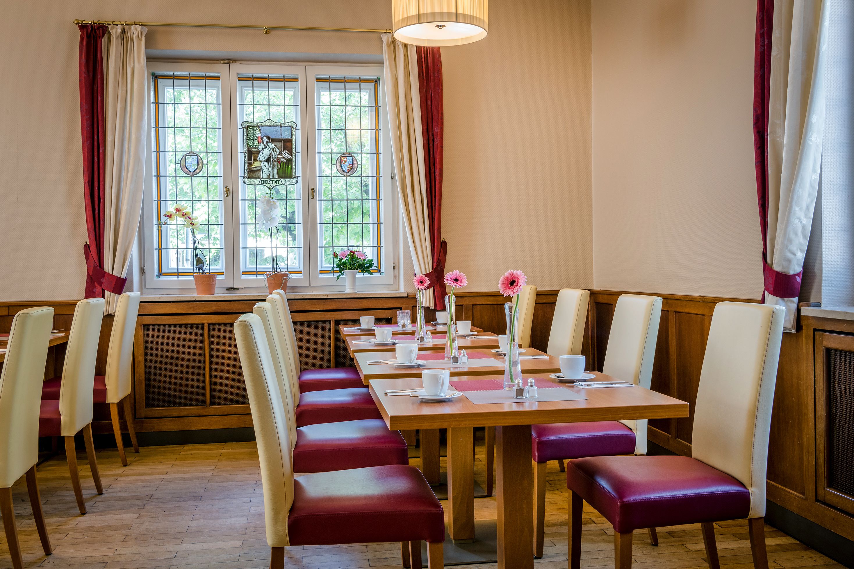 A dining area with wooden tables and red cushioned chairs, a window with curtains, and potted plants.