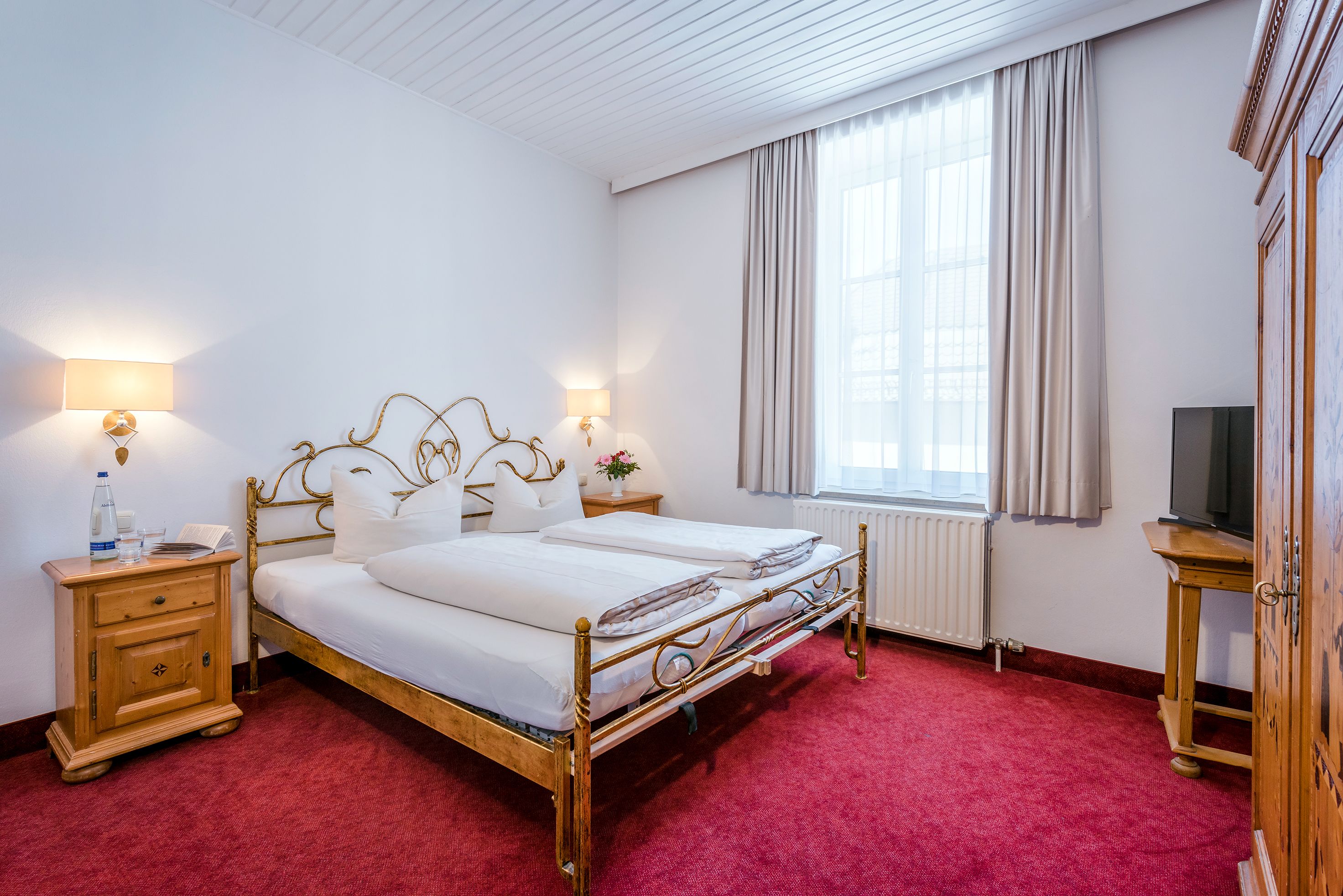 A double bed with a gold frame, two bedside tables with lamps, a wooden nightstand, a radiator, a television on a wooden stand, and a window with curtains.