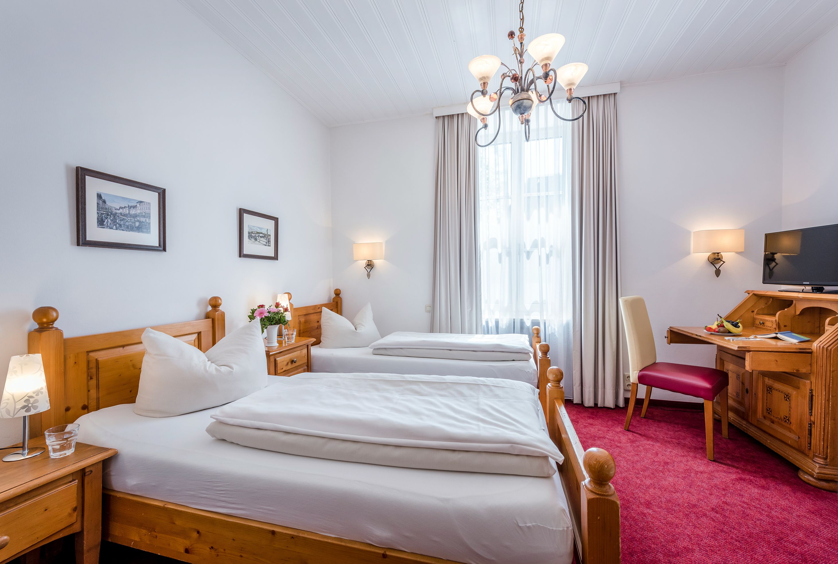 A hotel room with two beds, a desk, a chair, a TV, and a chandelier.