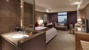 Habitación de hotel con una cama grande, un escritorio, un televisor, un baño con lavamanos y espejo, y vistas a la ciudad.