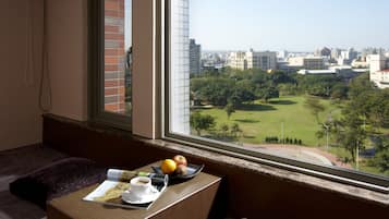 Una habitación con vistas a un paisaje urbano y a un parque. Hay una mesa con una taza, un plato de frutas y una revista.