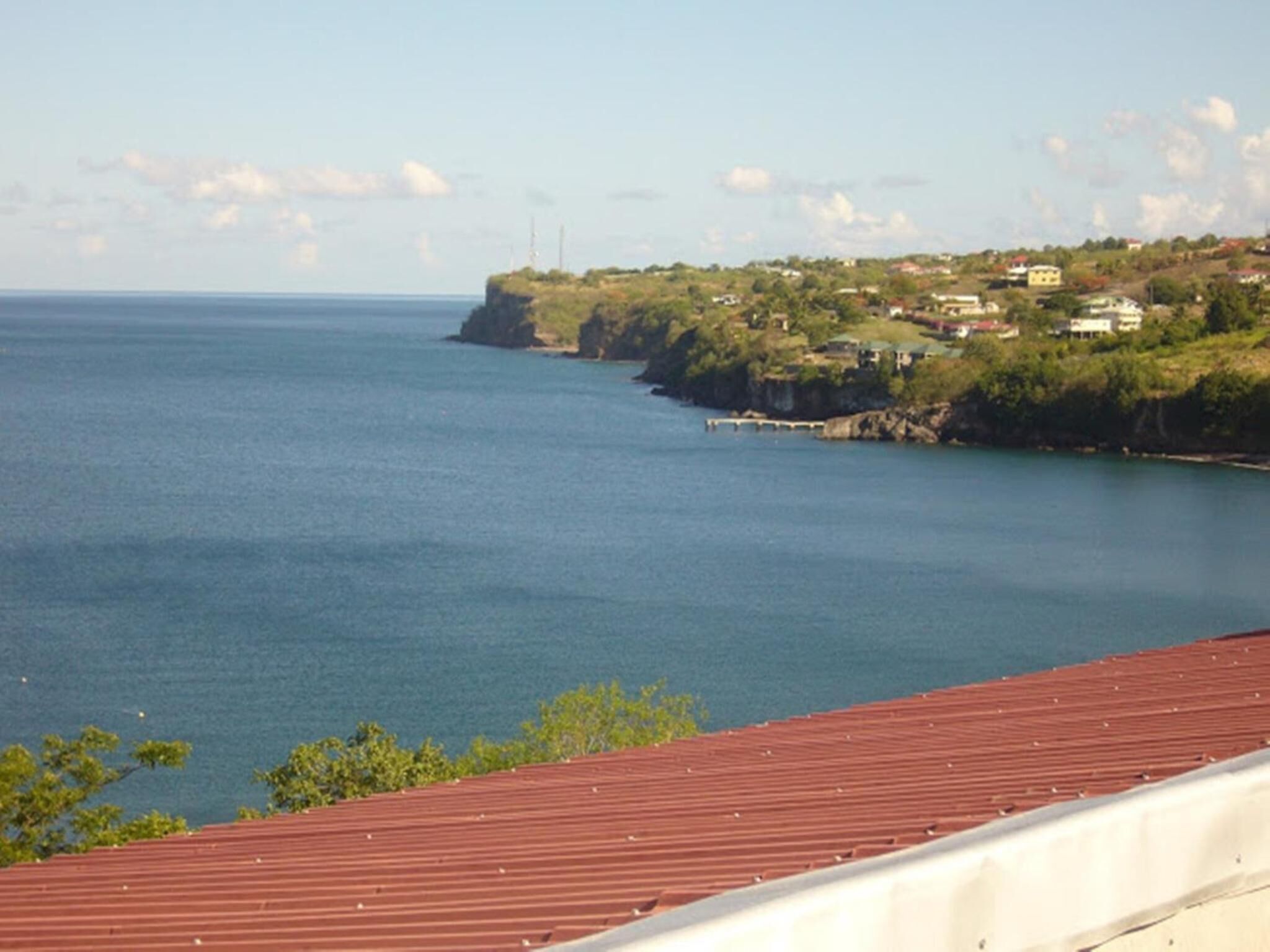 A coastal view with a red-roofed building, a clear blue sea, and a distant shoreline with houses and greenery.