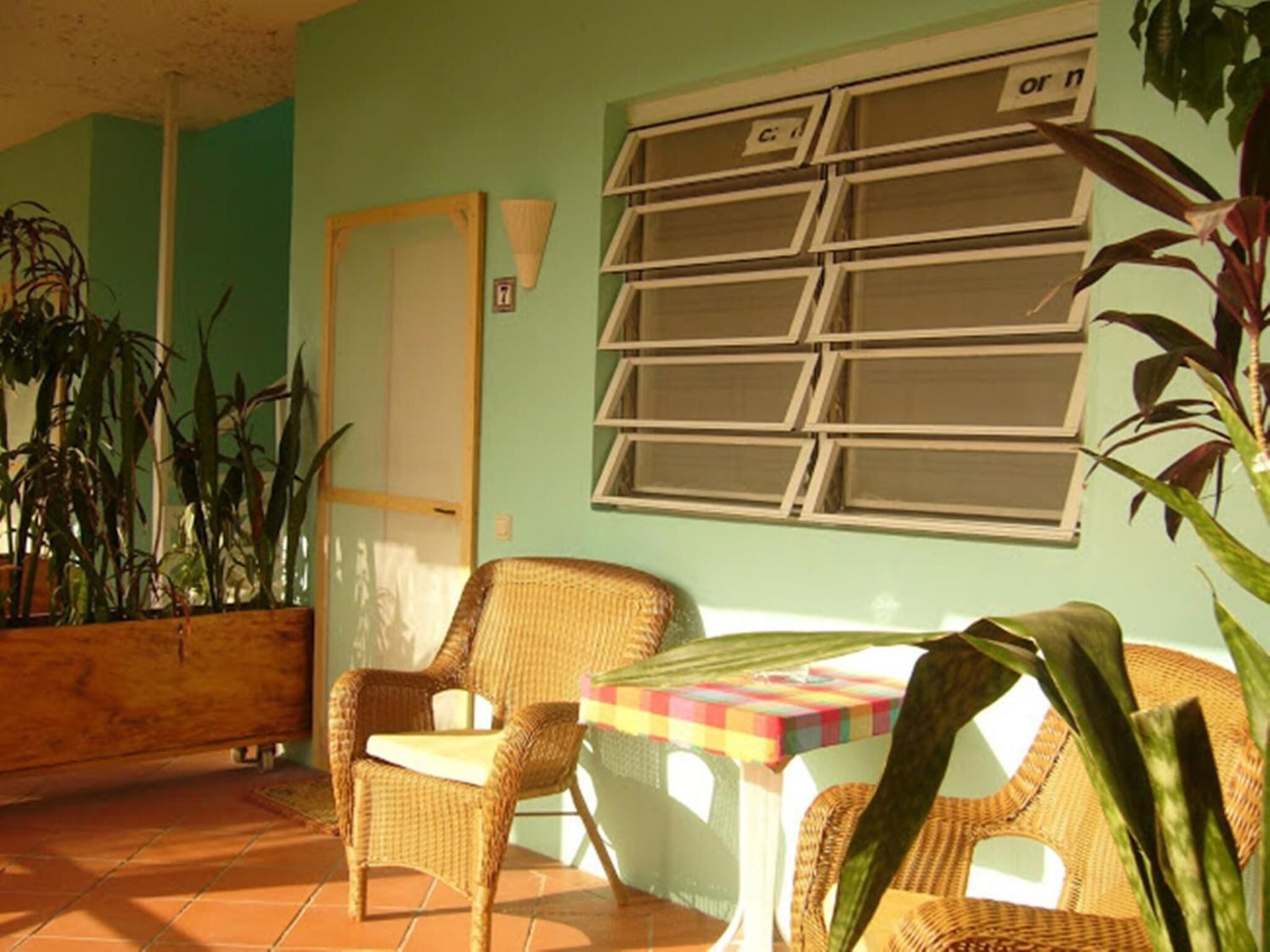 A room with a wicker chair, a small table with a colorful tablecloth, a window with blinds, and a potted plant.