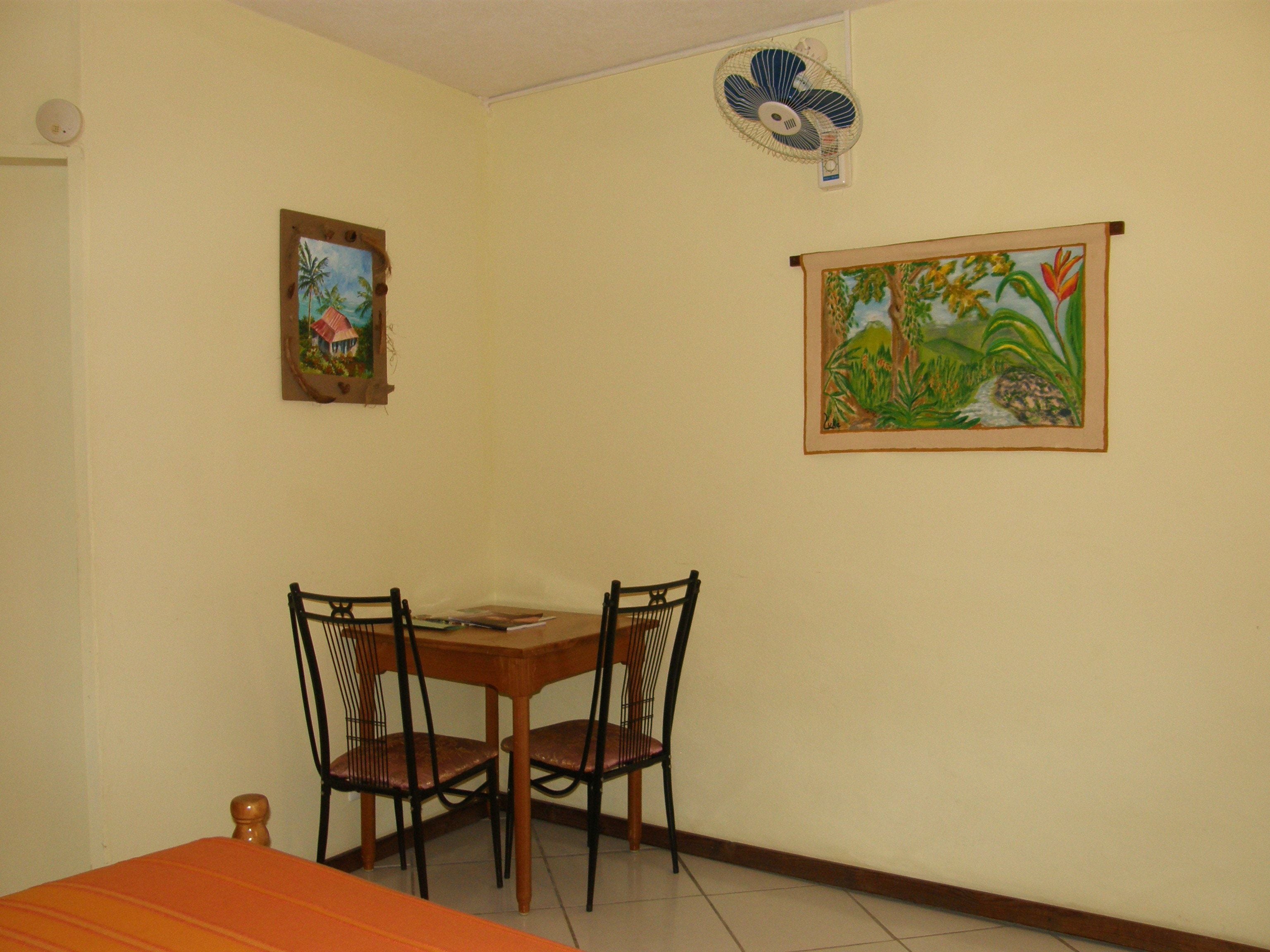 A dining area with a wooden table and chairs, two framed paintings on the wall, and a ceiling fan.
