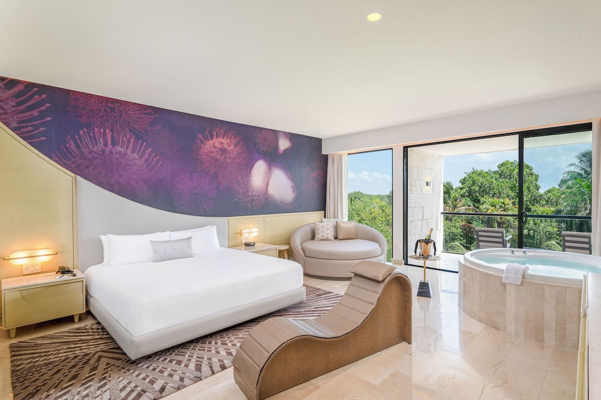 A modern hotel room with a large bed, a lounge chair, a jacuzzi, and a balcony with a view of greenery.