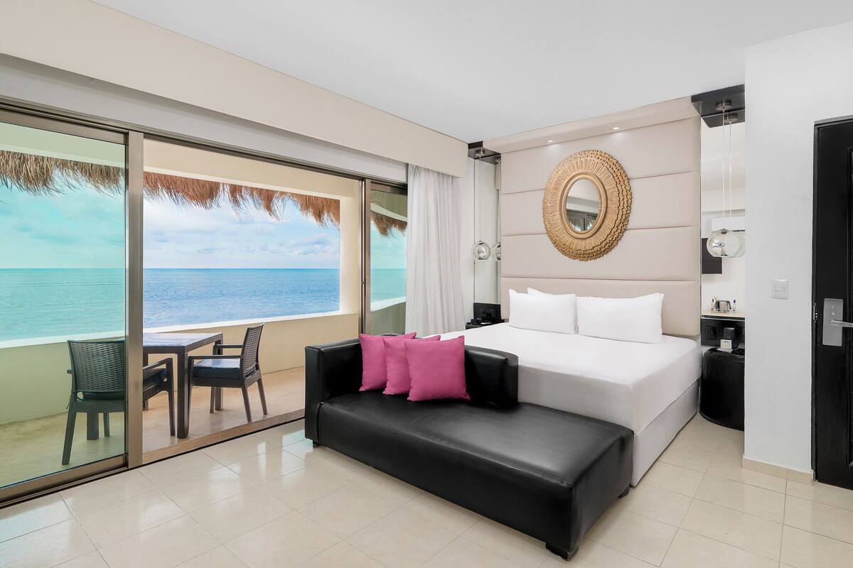 A modern hotel room with a large bed, a bench, a dining area, and a view of the sea.