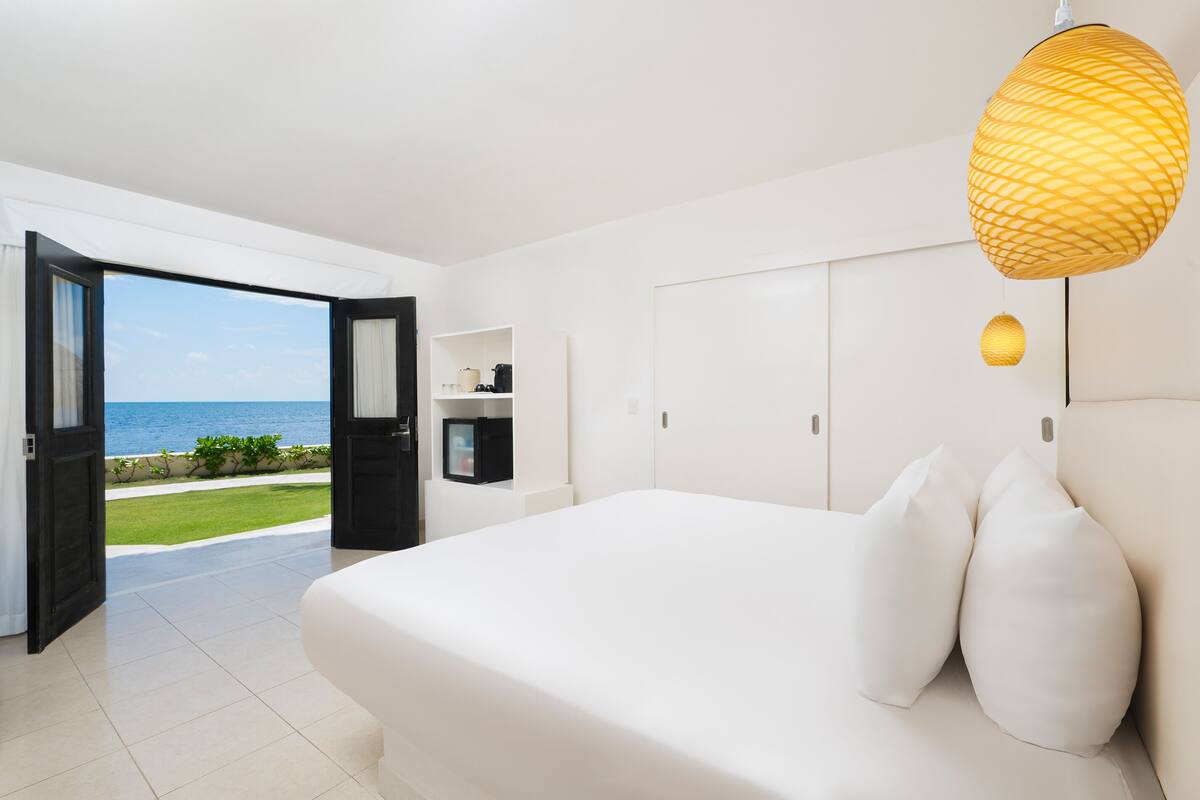 A bedroom with a large bed, white walls, a view of the sea through an open door, and a hanging light fixture.