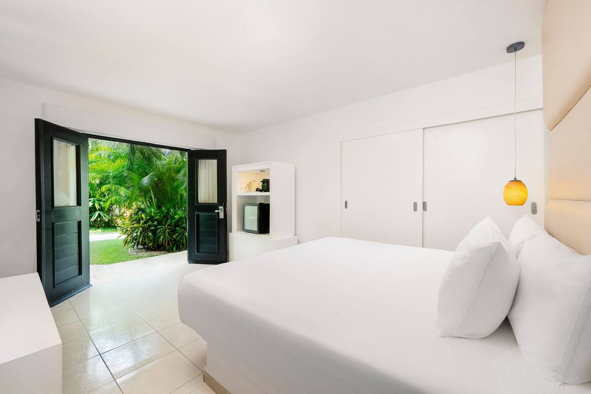 A modern bedroom with a large bed, white bedding, a bedside table, a wardrobe, and a view of greenery outside.