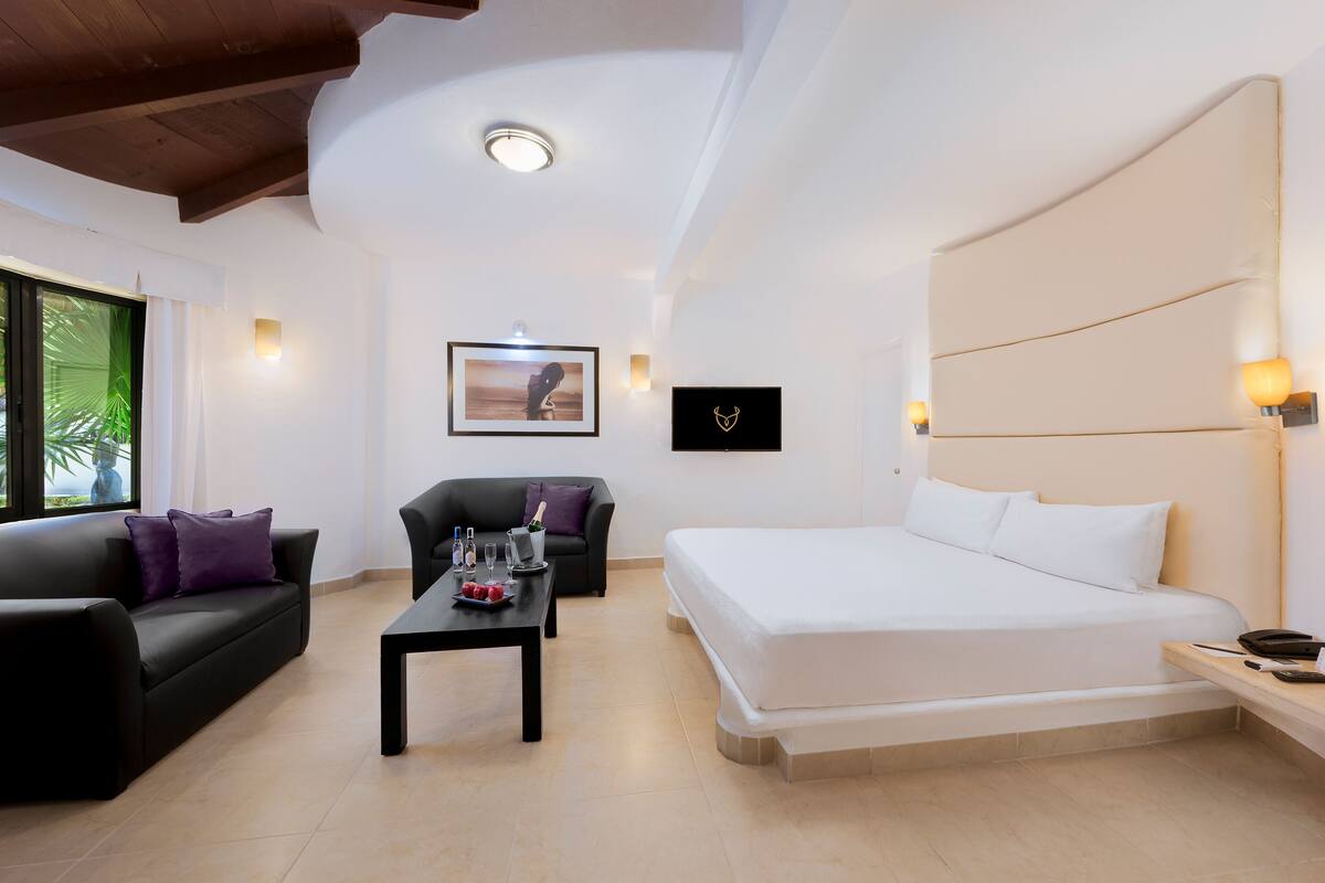 A modern hotel room with a large bed, a sofa, a coffee table, and a desk.