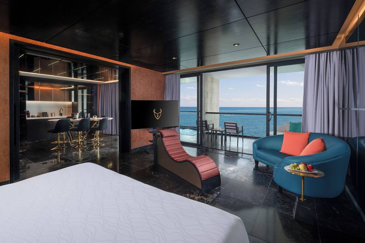 A modern hotel room with a large bed, a massage chair, a sofa, and a dining area with a view of the sea.