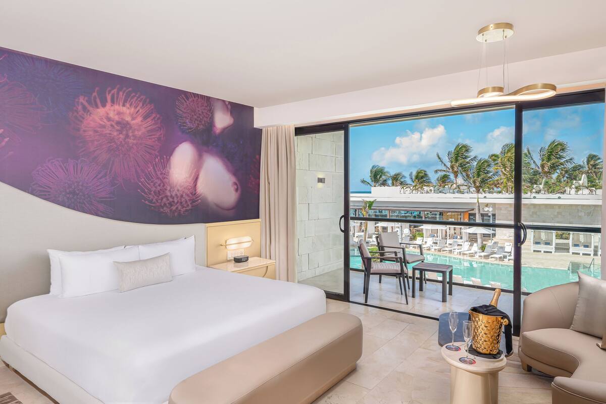 A hotel room with a large bed, a balcony with a pool view, and a dining area.