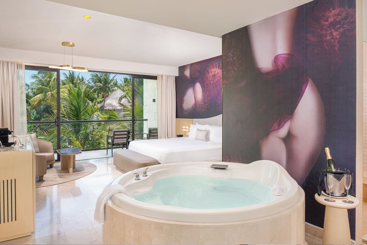 A modern hotel room with a large bed, a jacuzzi, a balcony with a view of palm trees, and a large artwork on the wall.