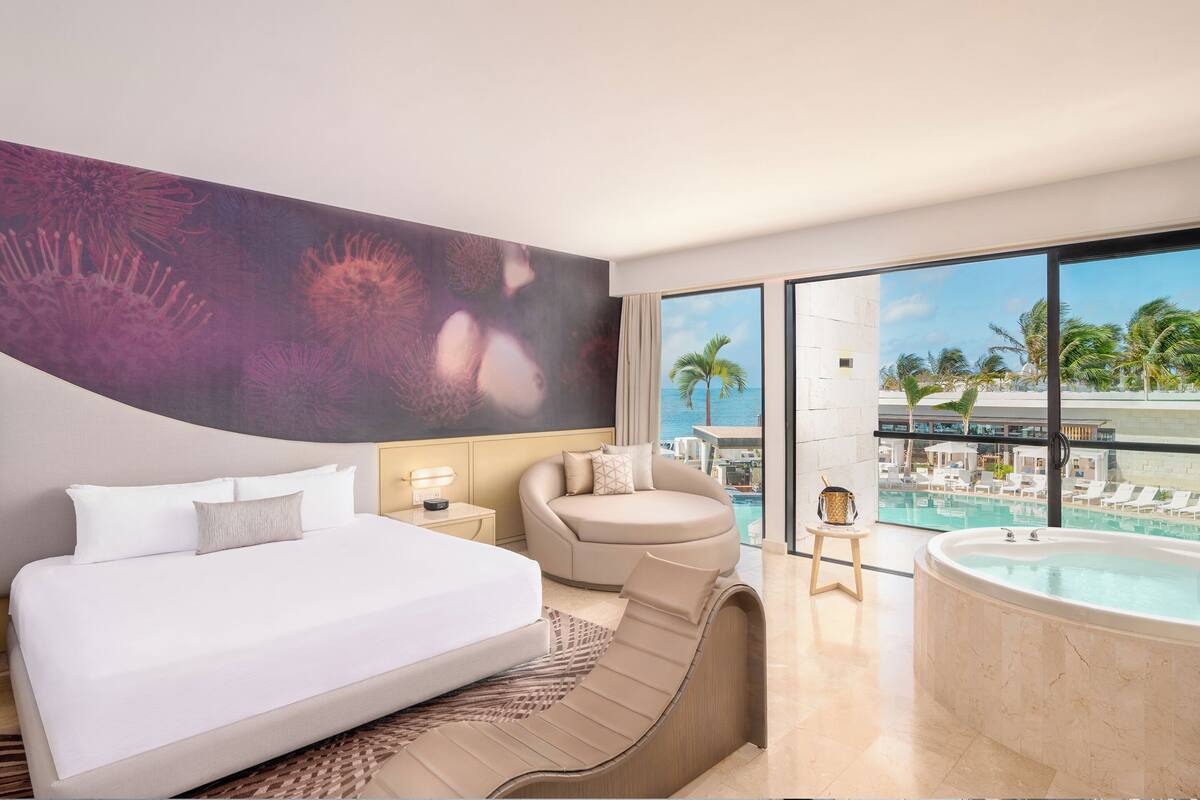 A hotel room with a bed, a sofa, a bathtub, and a large window with a view of a pool and palm trees.
