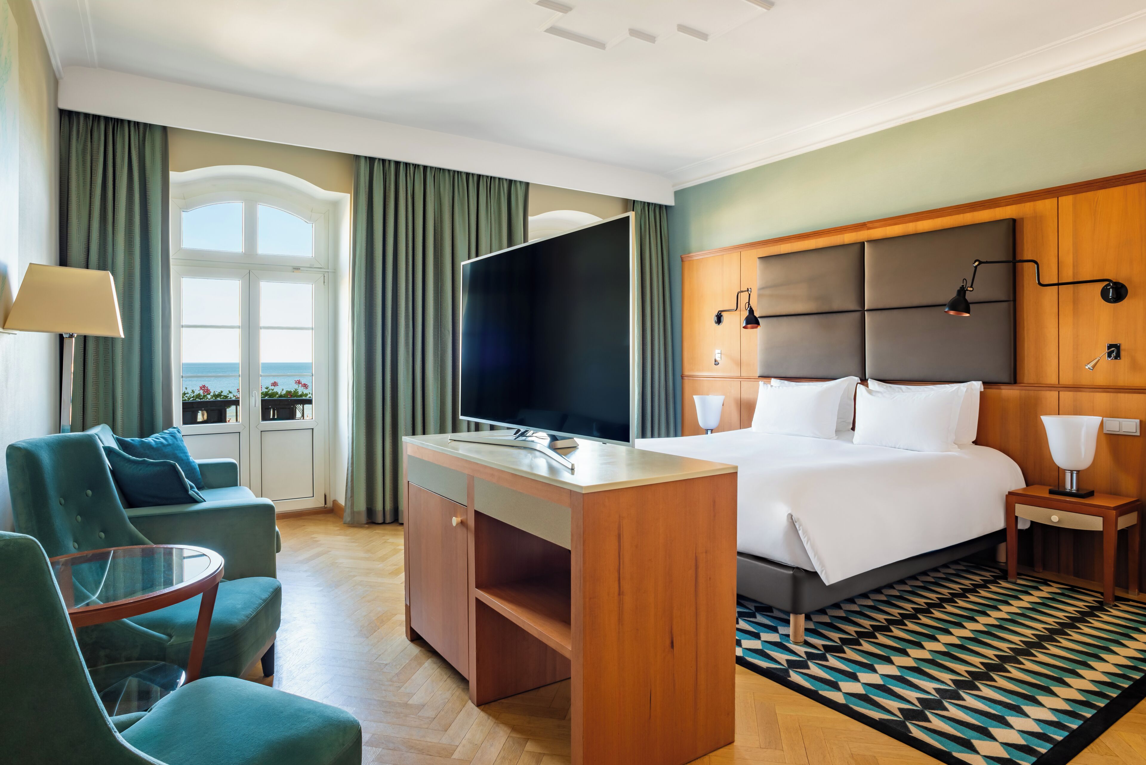 Ett hotellrum med en säng, ett skrivbord med en tv, en sittgrupp med stolar och utsikt över havet genom ett fönster.