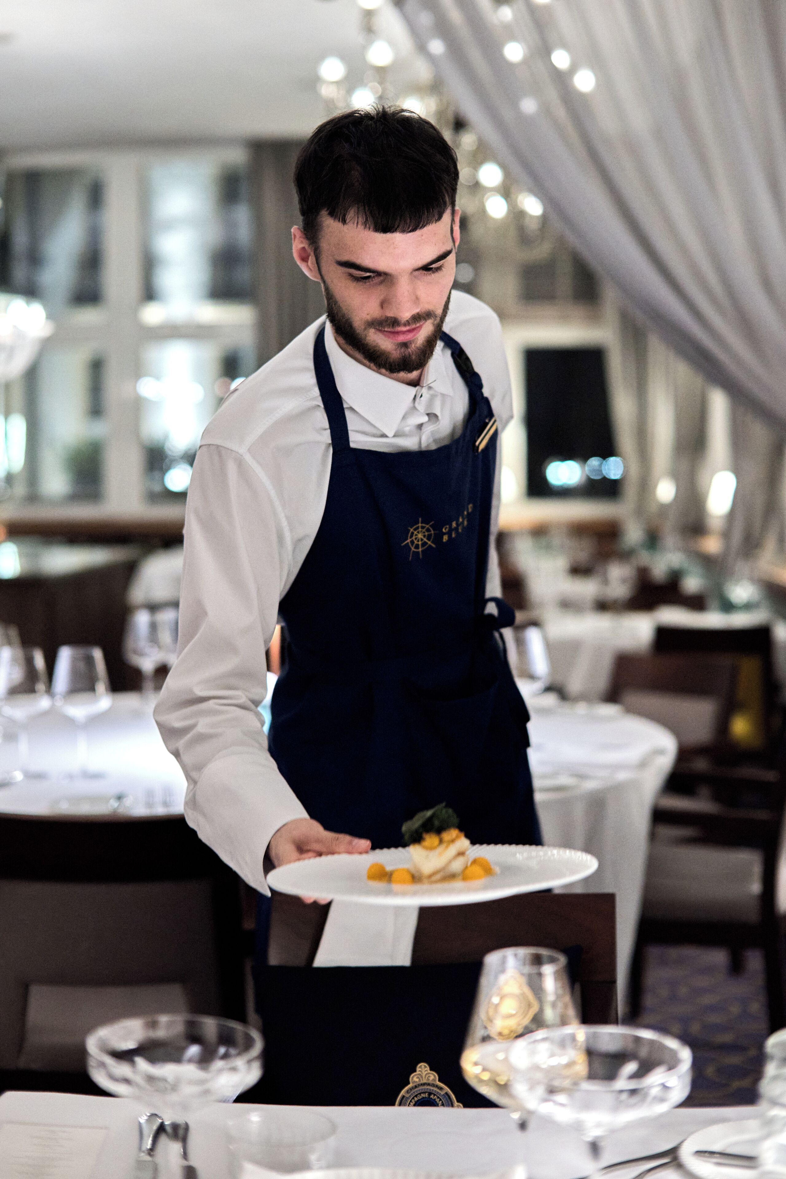 Un camarero con uniforme elegante sirve un plato en un comedor bien iluminado y decorado con esmero, con mesas dispuestas para comer.