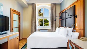 Habitación de hotel con cama, escritorio, televisión, ventana con cortinas y vistas a árboles.