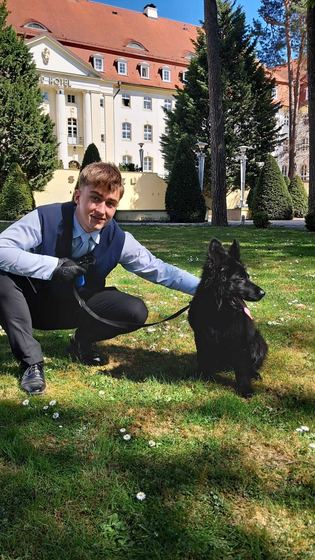 En person hukar på gräset tillsammans med en svart hund, och i bakgrunden syns en byggnad med ett rött tak.