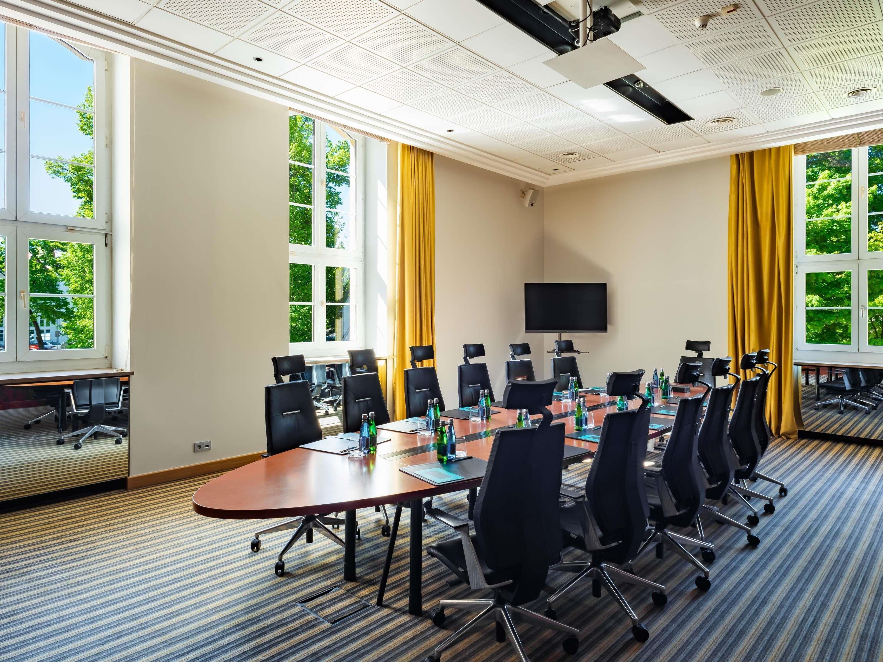 Ett konferensrum med ett stort bord, stolar, en platt-TV och fönster med gardiner.