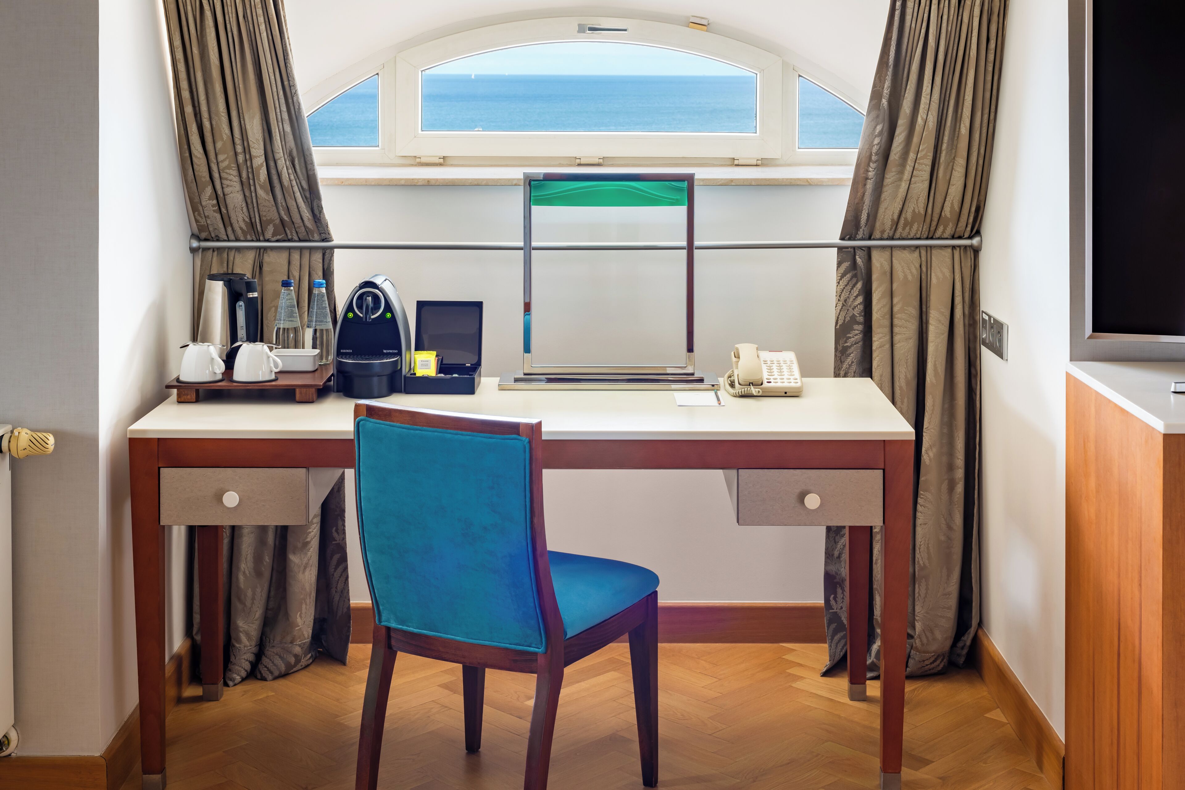 A room with a desk, chair, window, curtains, and a view of the sea.