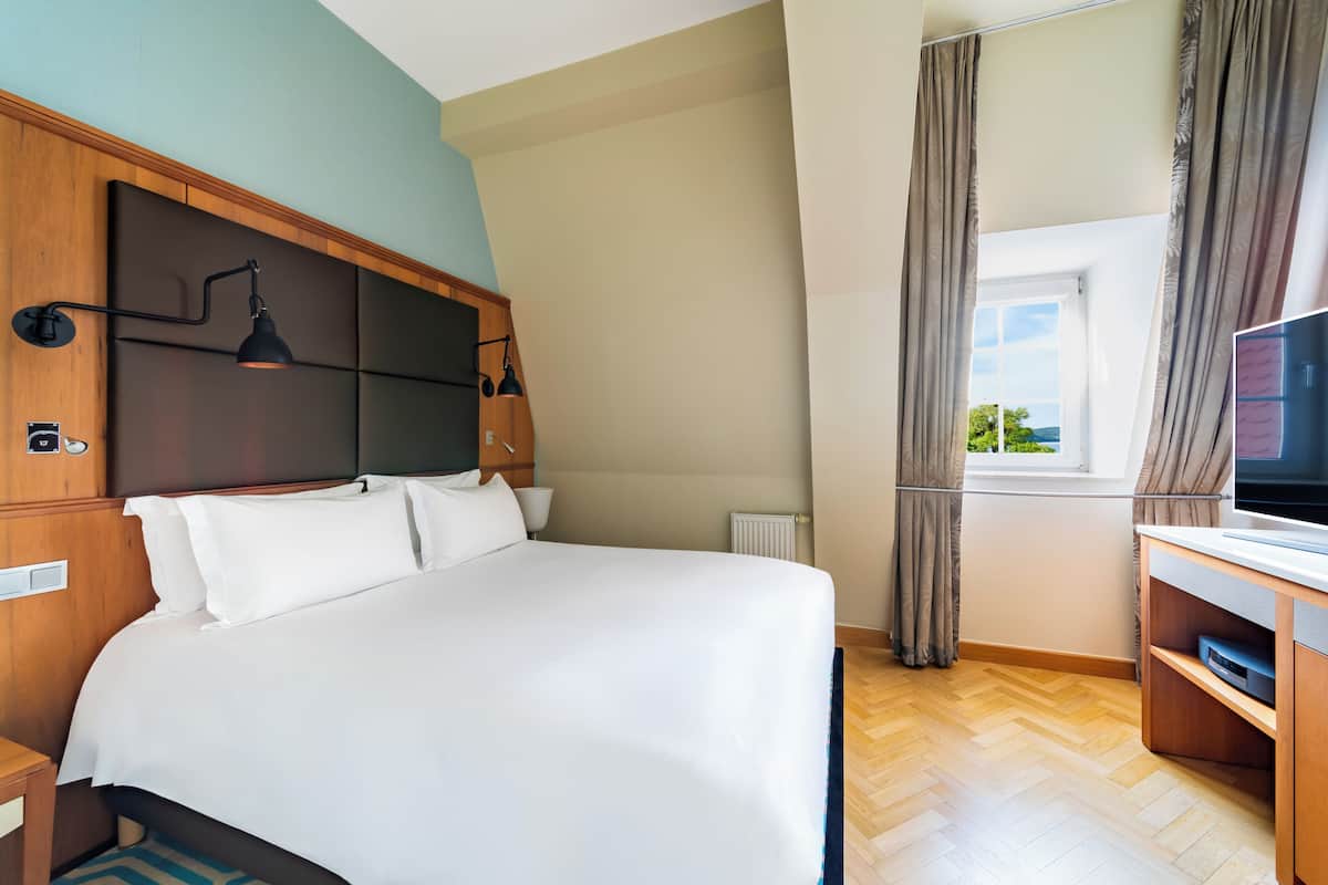 Hotellihuone, jossa on sänky, televisio, ikkuna, jossa on verhot, ja puinen pääty.