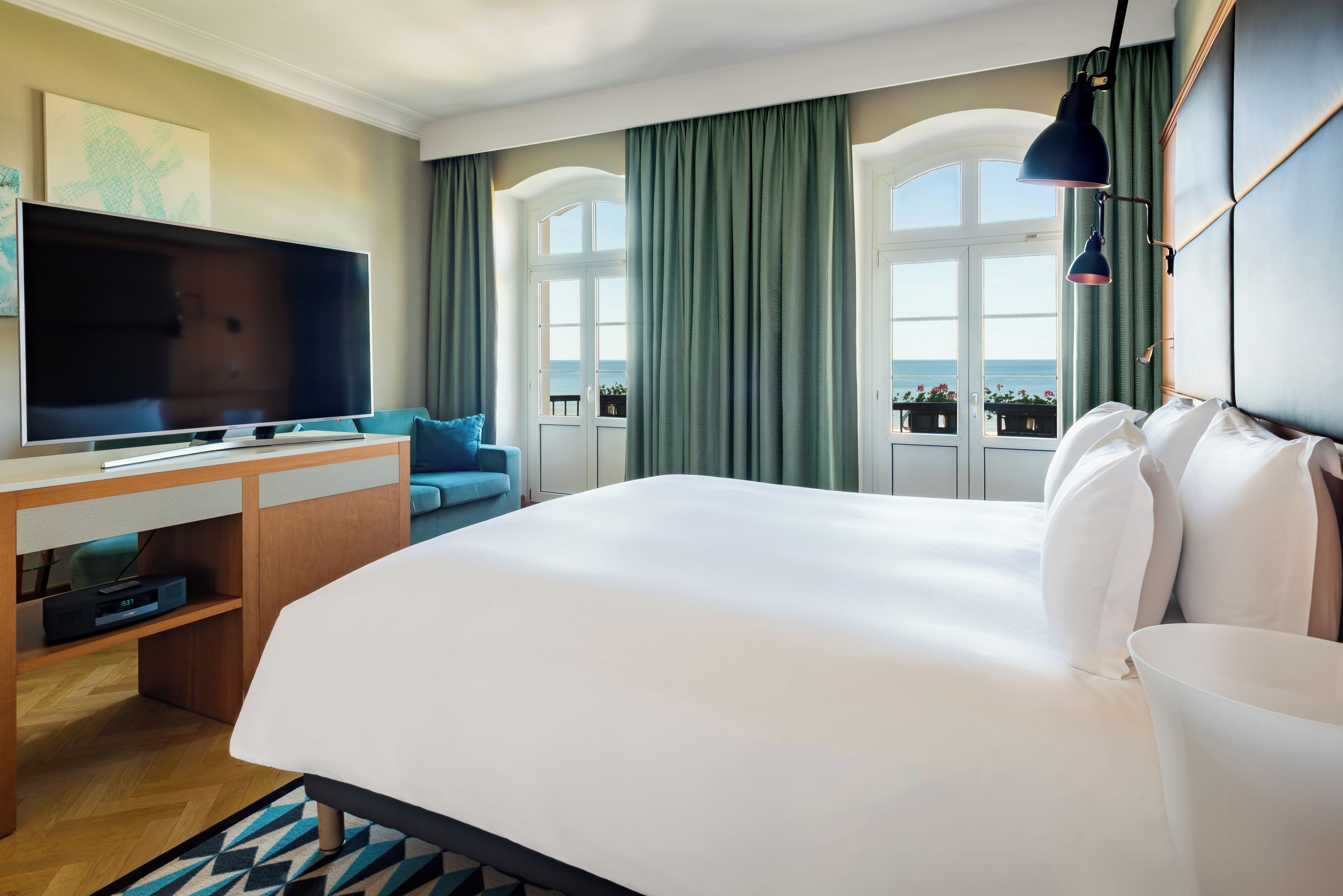 A hotel room with a large bed, a desk with a flat-screen TV, a view of the sea through large windows, and a balcony with plants.