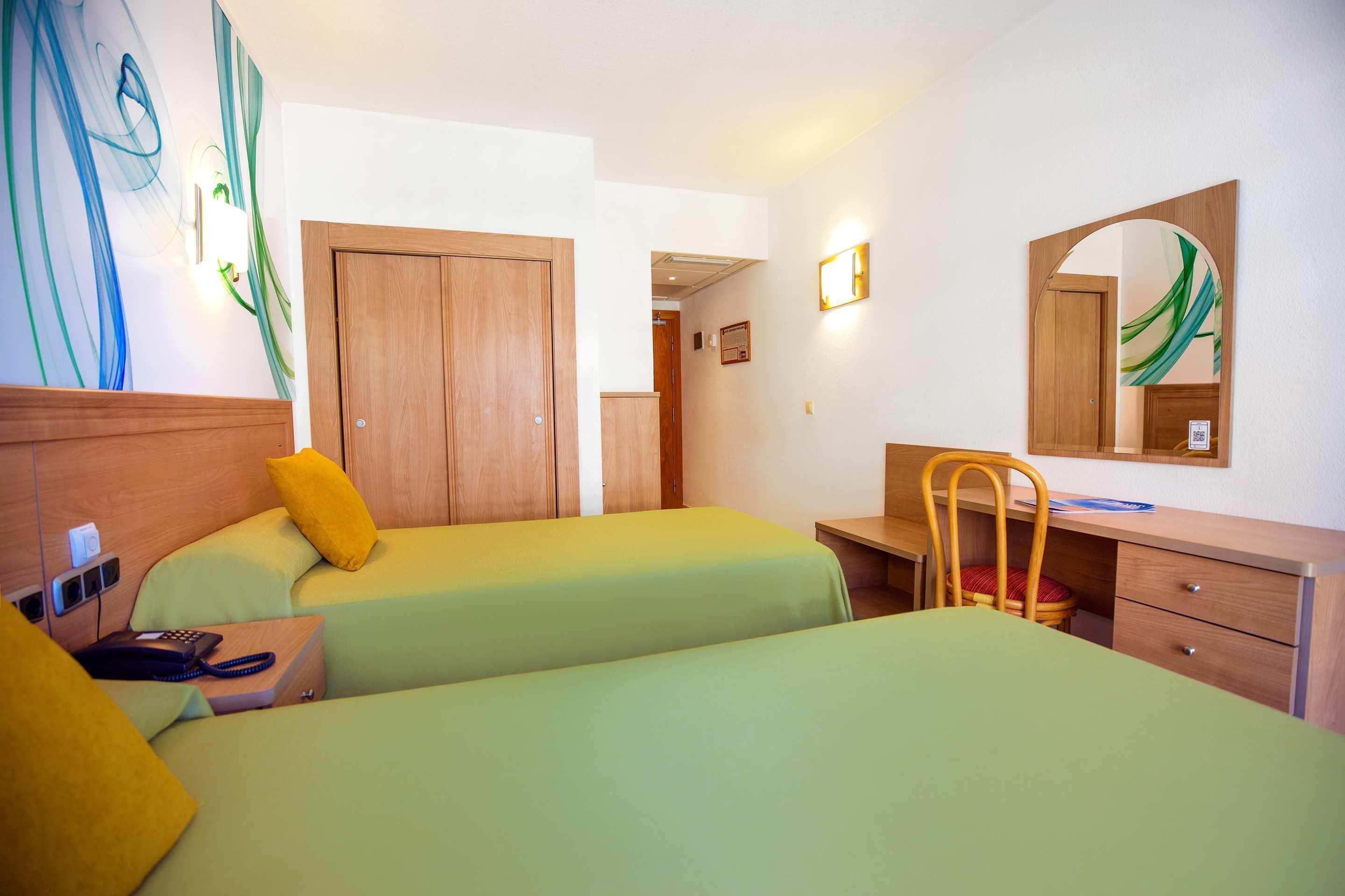 A hotel room with two beds, a desk, a chair, a wooden wardrobe, and a mirror.