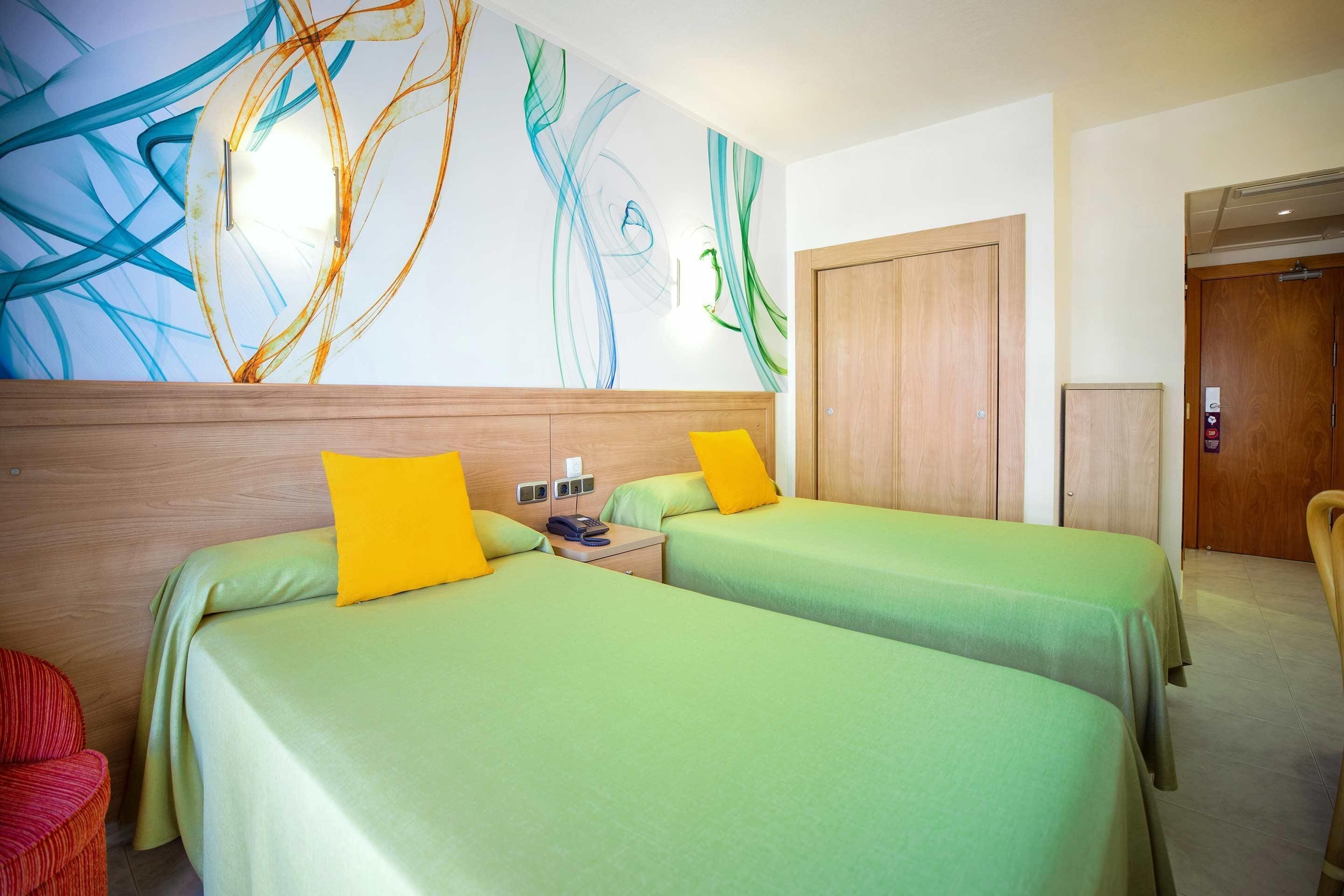 Quarto de hotel com duas camas, cabeceira de madeira, parede com arte abstrata, guarda-roupa de madeira e uma cadeira vermelha.