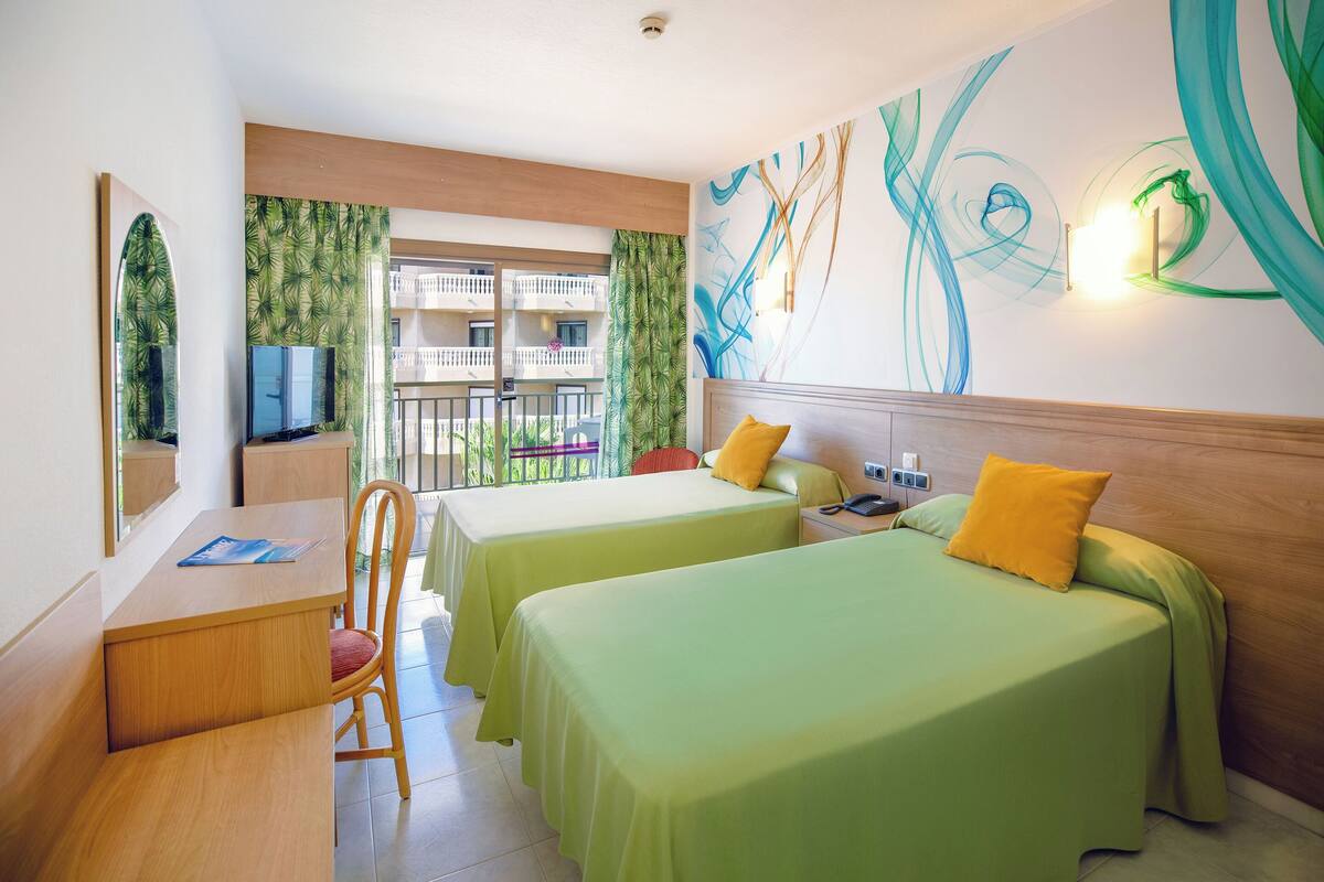 Hotellihuone, jossa on kaksi sänkyä, työpöytä, tuoli ja parveke, jolta on näkymä muihin rakennuksiin.