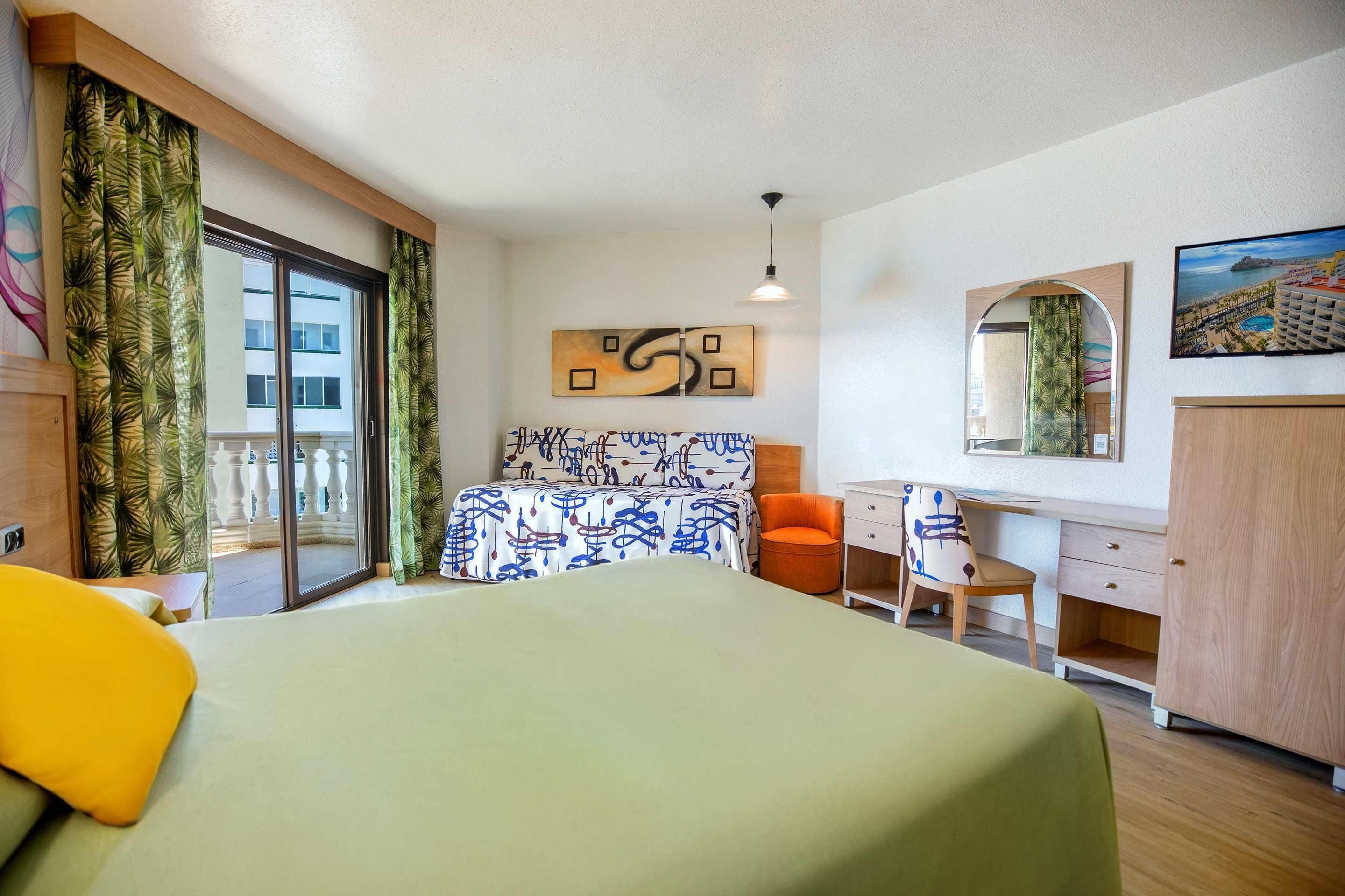 A hotel room with a bed, a desk, a chair, a wardrobe, a large window with curtains, and artwork on the walls.