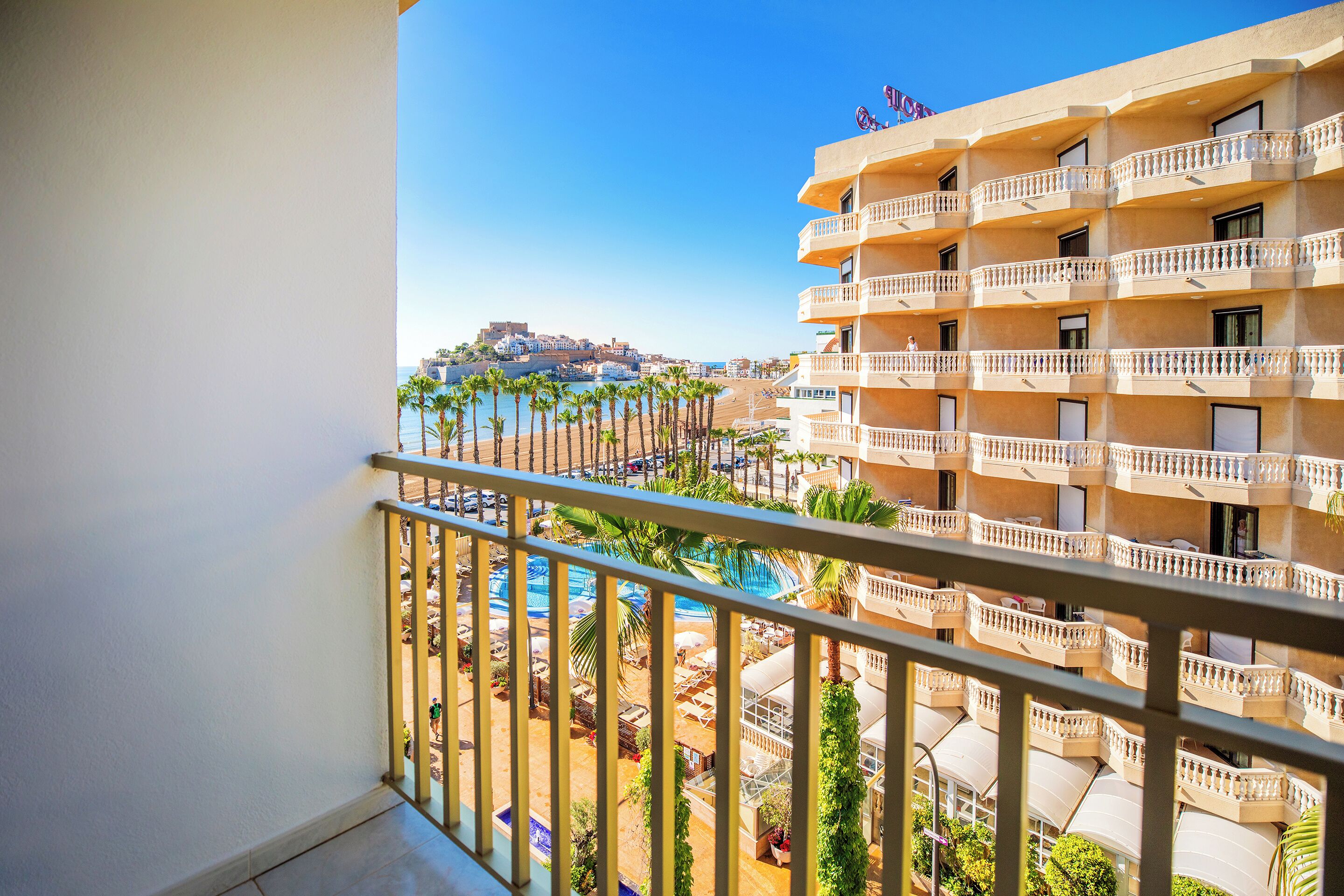 A hotel balcony with a view of a pool, palm trees, and a distant cityscape.